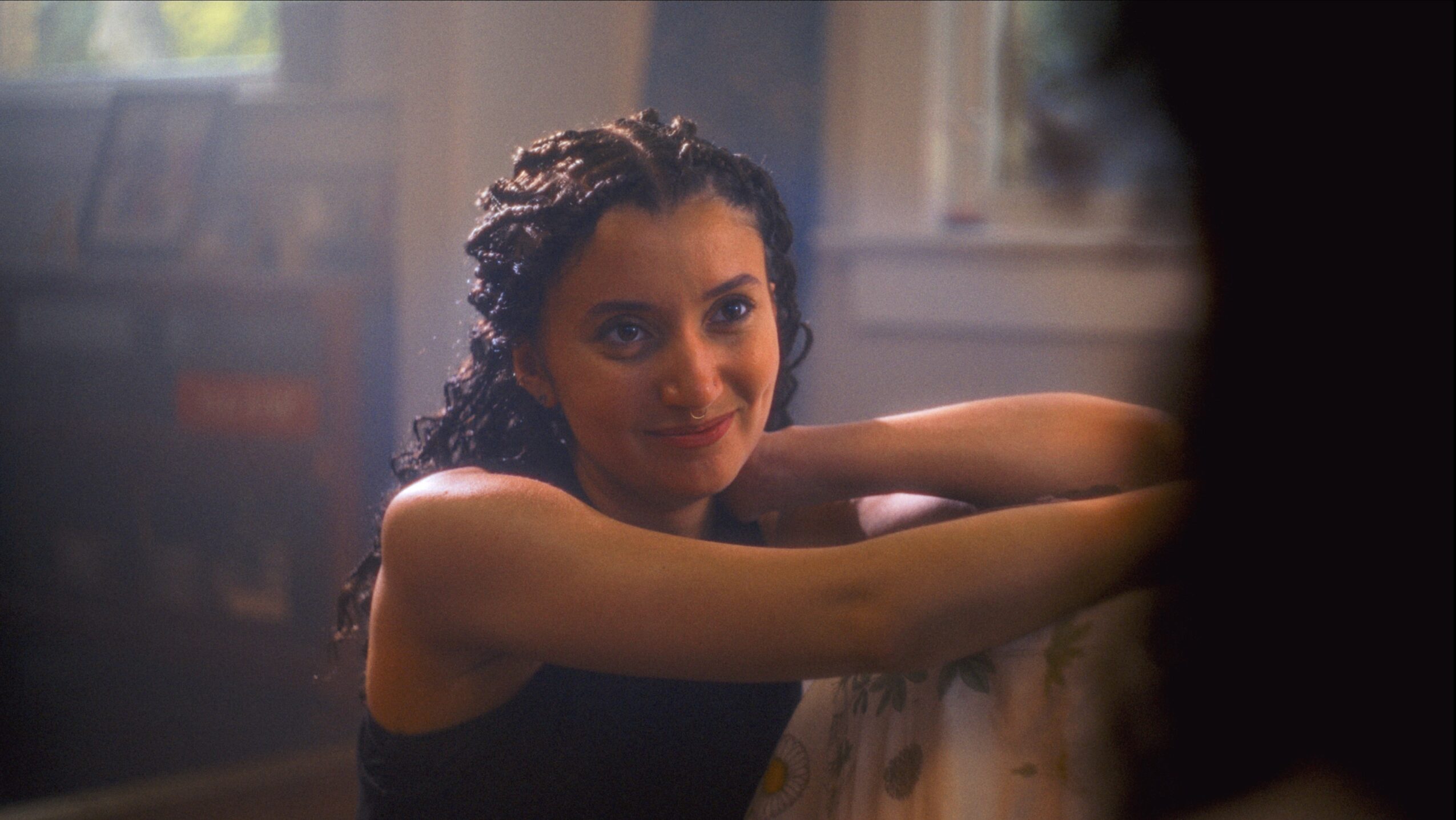 Woman with dark curly hair smiling, resting her arms on a surface indoors.