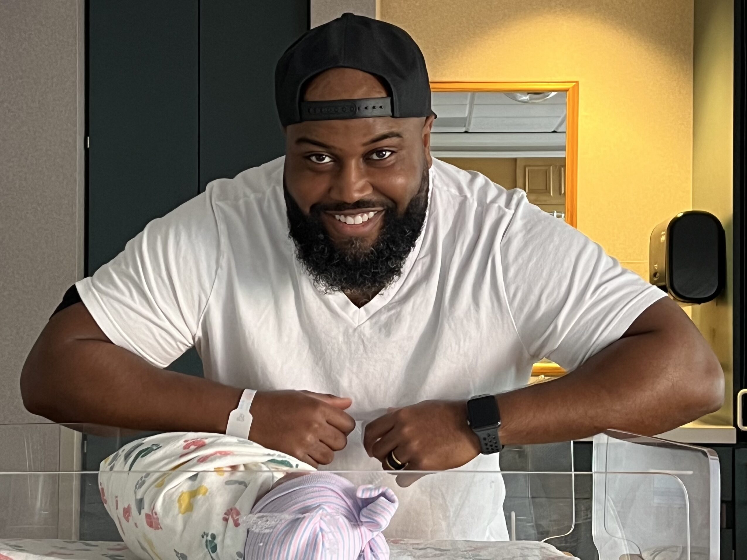 Man with a beard wearing a black cap backwards and a white t-shirt, smiling, leaning on a counter.