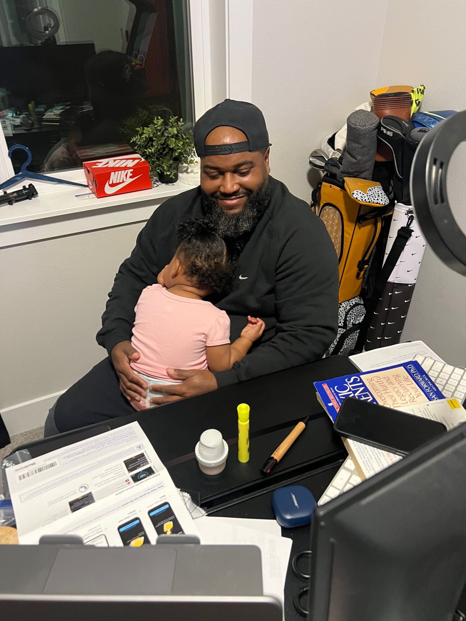 Person in black jacket and glasses holding a pregnant woman in pink shirt, sitting at a desk with papers and office supplies.