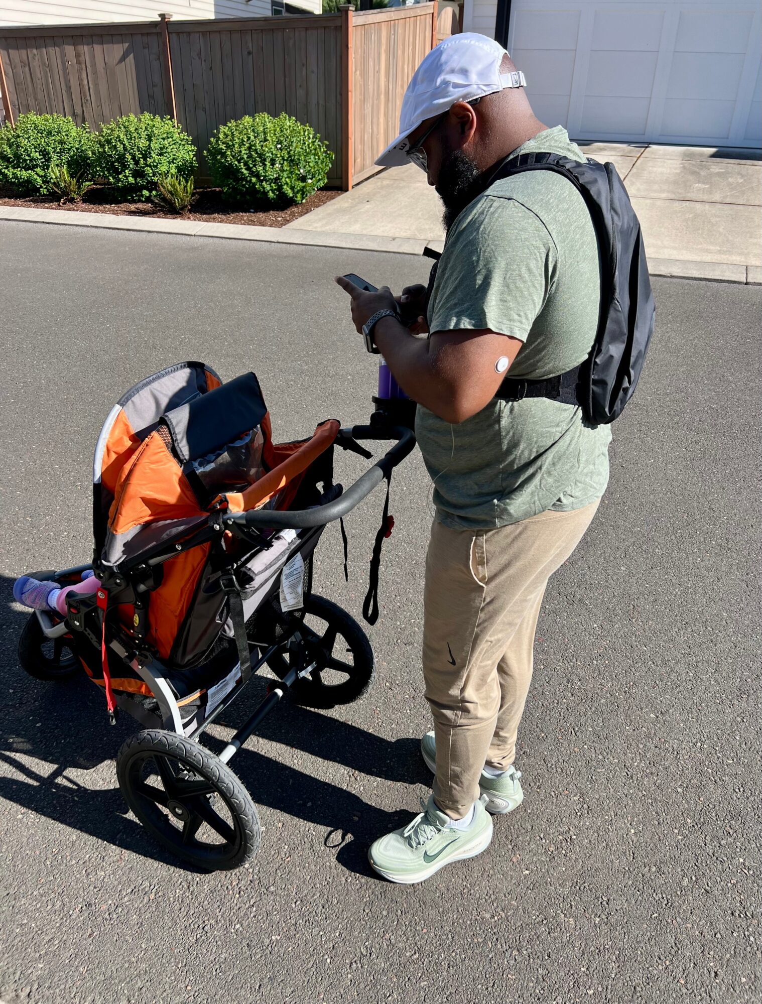Man with a backpack looking at his phone next to a stroller on a paved street, with a fence and bushes in background.