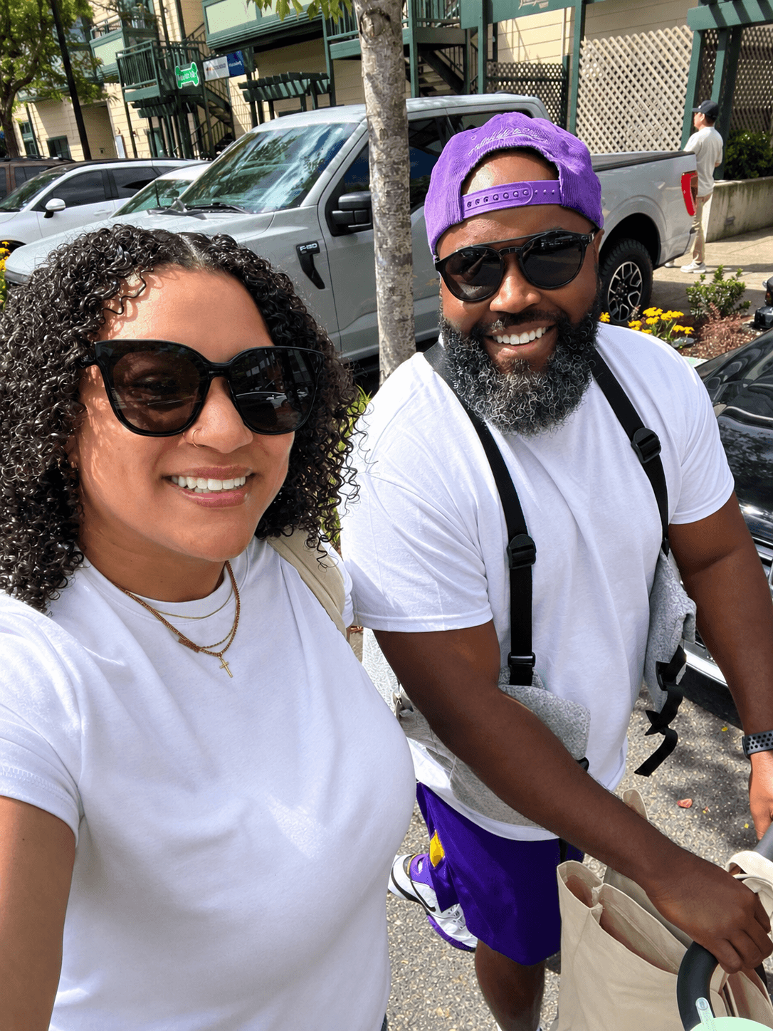 Two smiling people wearing sunglasses outdoors on a sunny day, with cars and trees in the background.