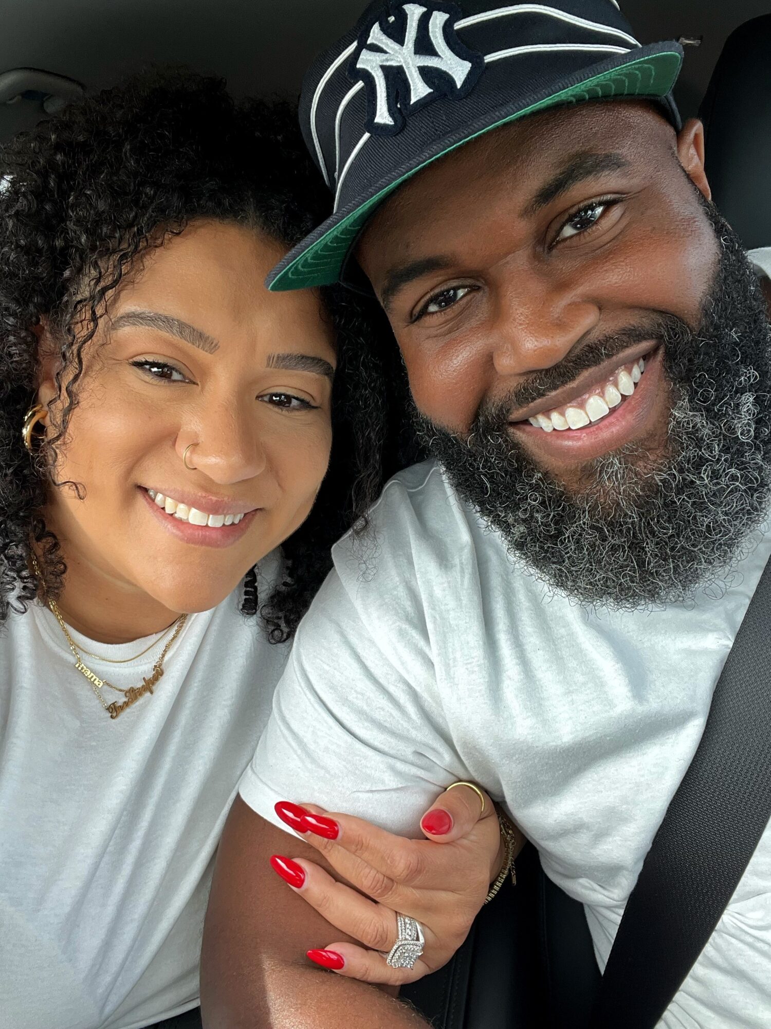 Two smiling people, a woman with curly hair and a man with a beard wearing a New York Yankees cap, sitting close together.