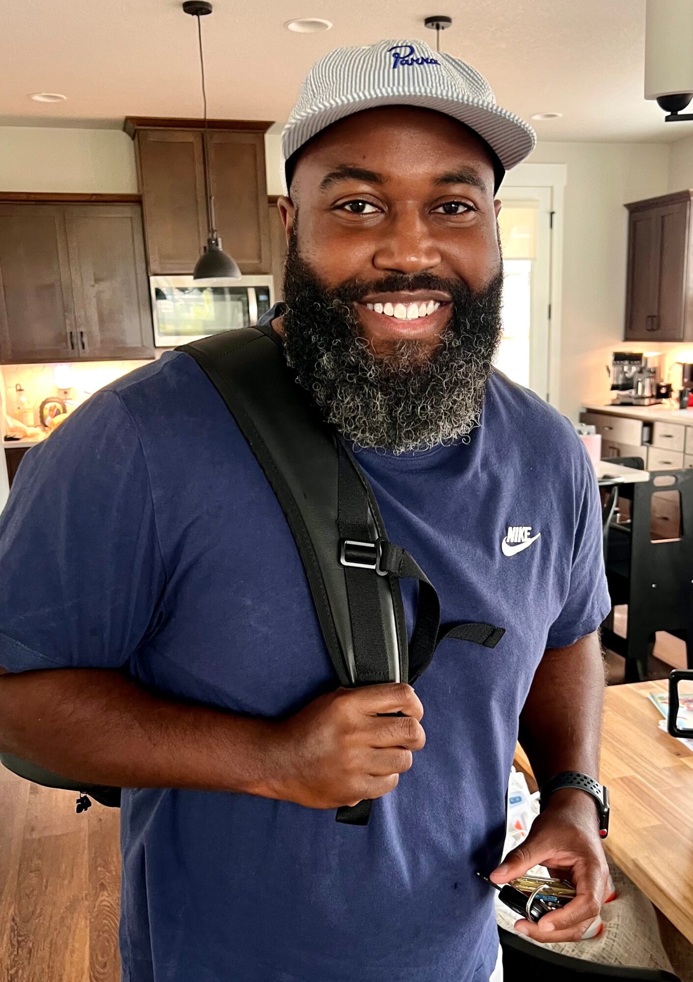 Smiling man with a beard wearing a white cap and blue Nike t-shirt, carrying a backpack in a kitchen.