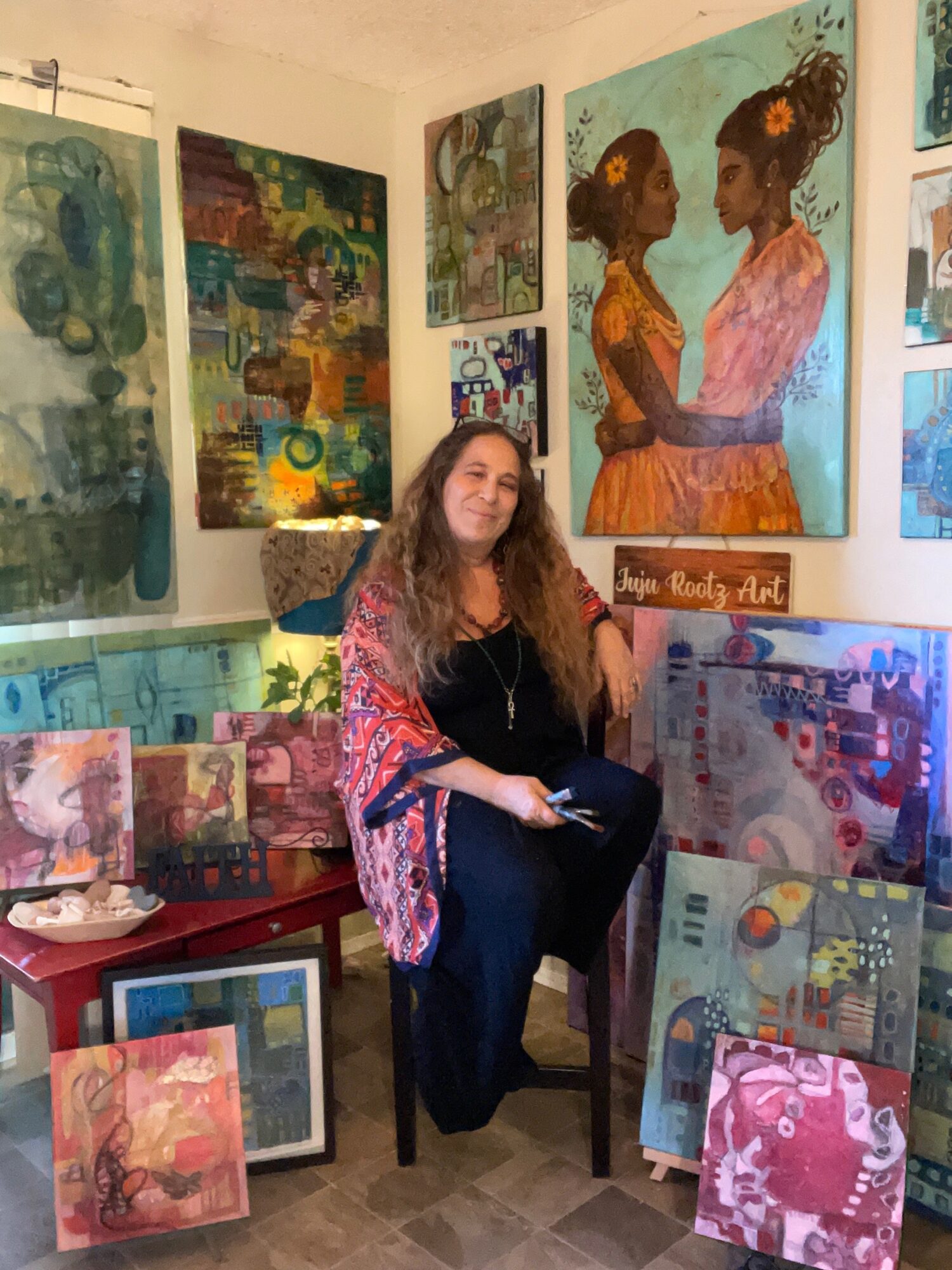 Woman with long hair sitting in an art gallery surrounded by colorful paintings on walls and floor.
