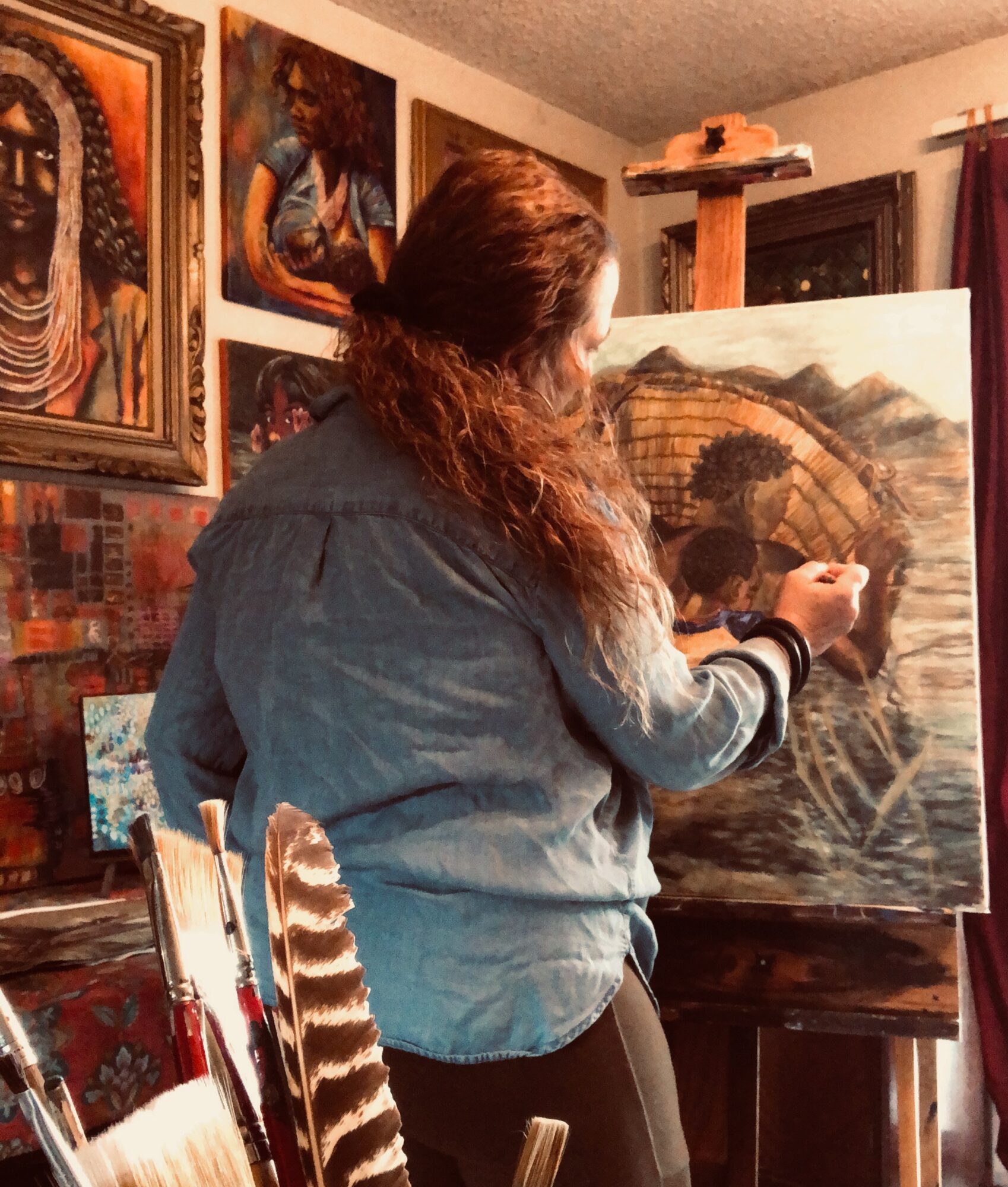 Person with long hair painting a landscape on canvas in an art studio, surrounded by artwork and supplies.