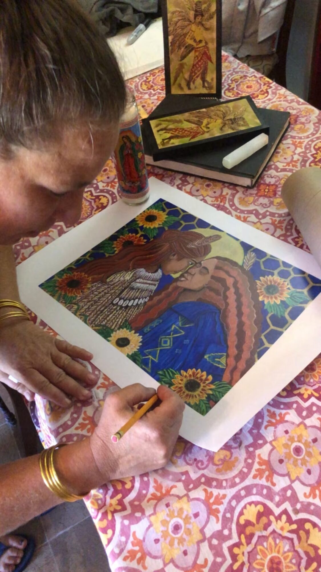 Person working on a colorful artwork of a woman in traditional attire, surrounded by flowers, on a table with art supplies.