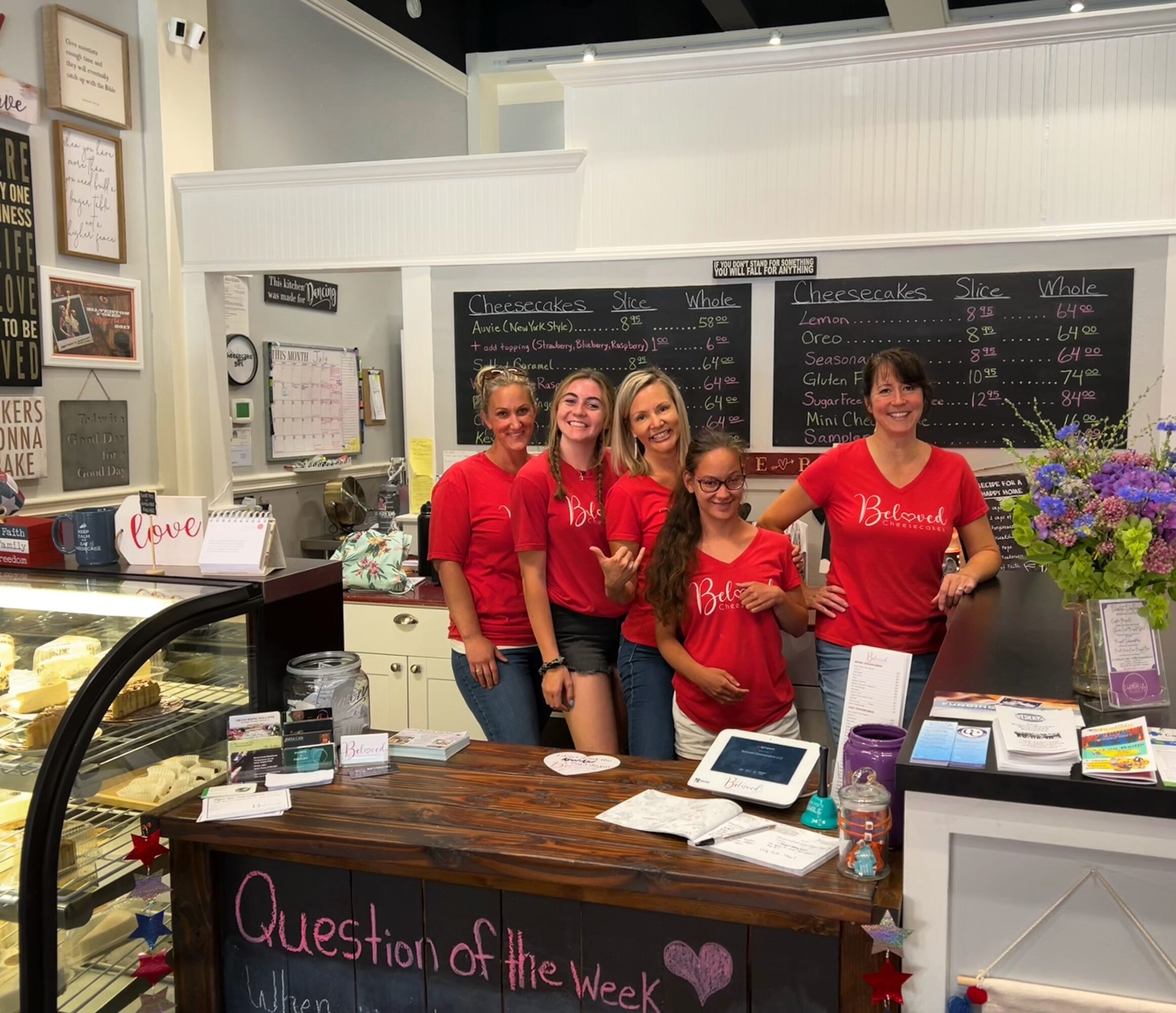 Four women in red shirts stand behind a counter in a cafe, smiling, with a chalkboard menu behind them and flowers on the side.