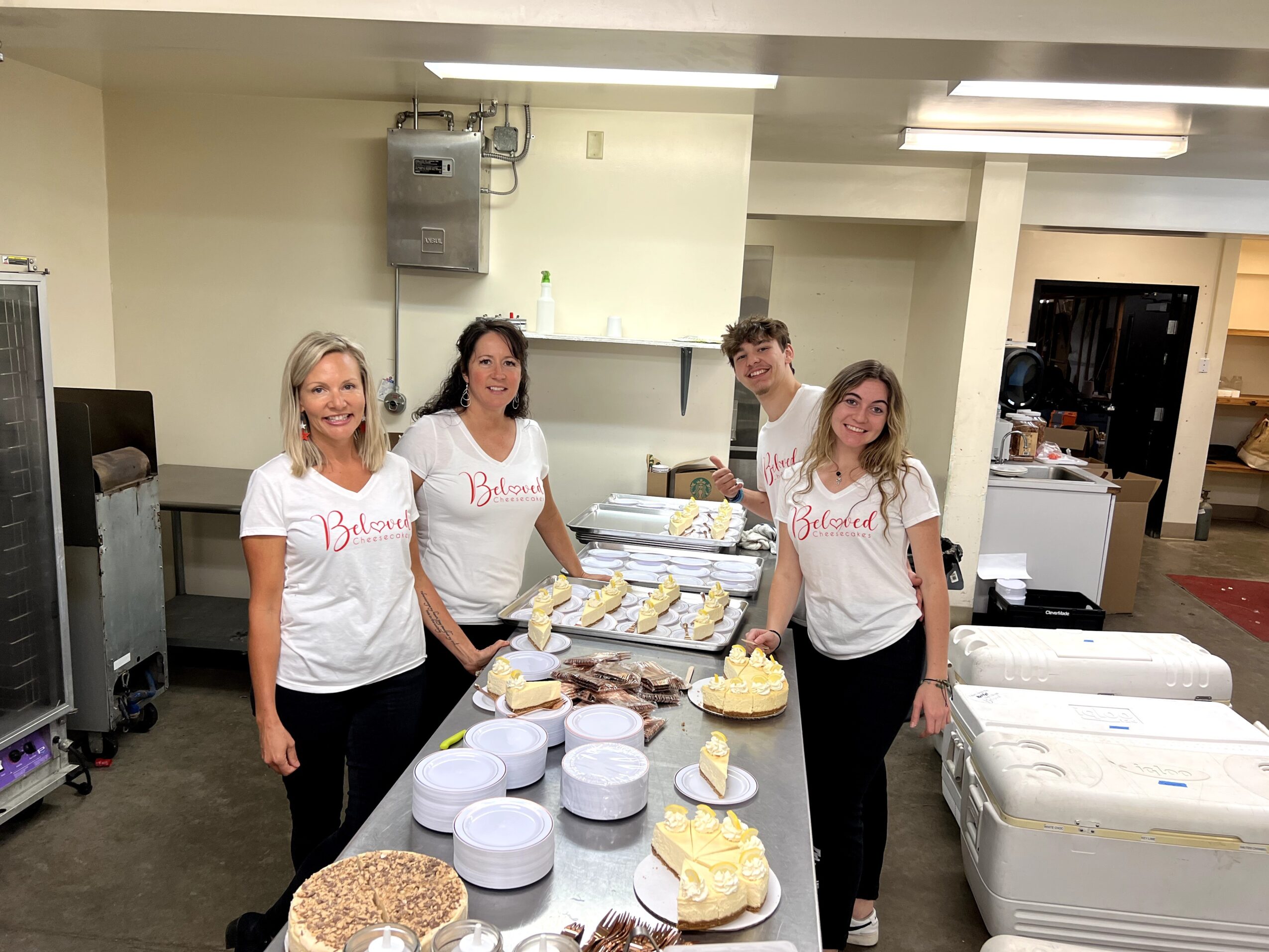Four women stand behind a table with cakes and desserts in a room, smiling at the camera.