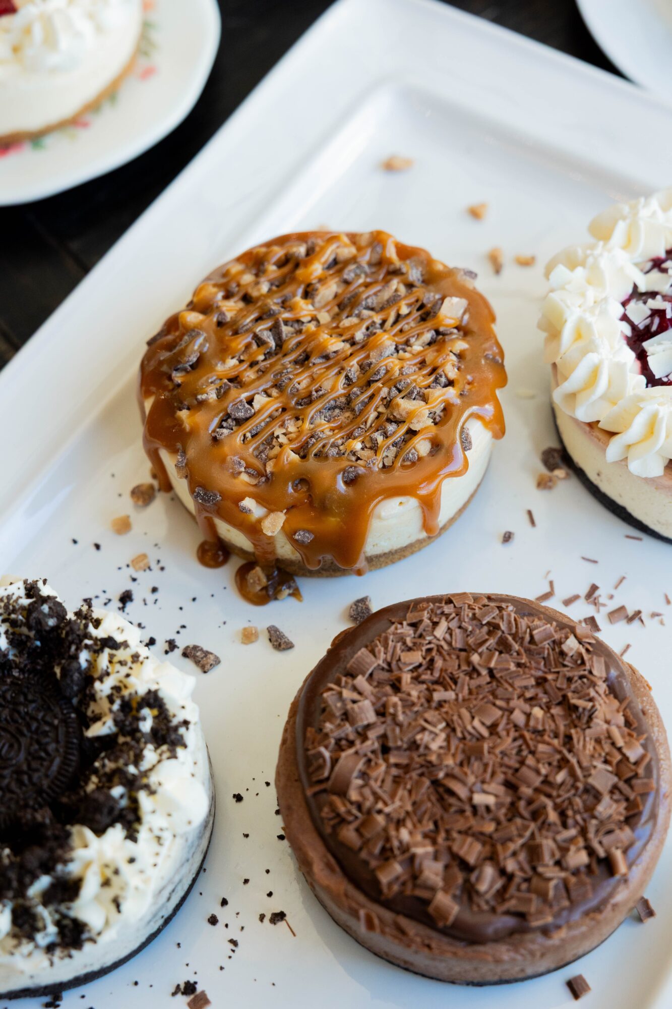 Close-up of four assorted mini cheesecakes on a white rectangular plate, with caramel, chocolate, and toppings, on a dark surface.