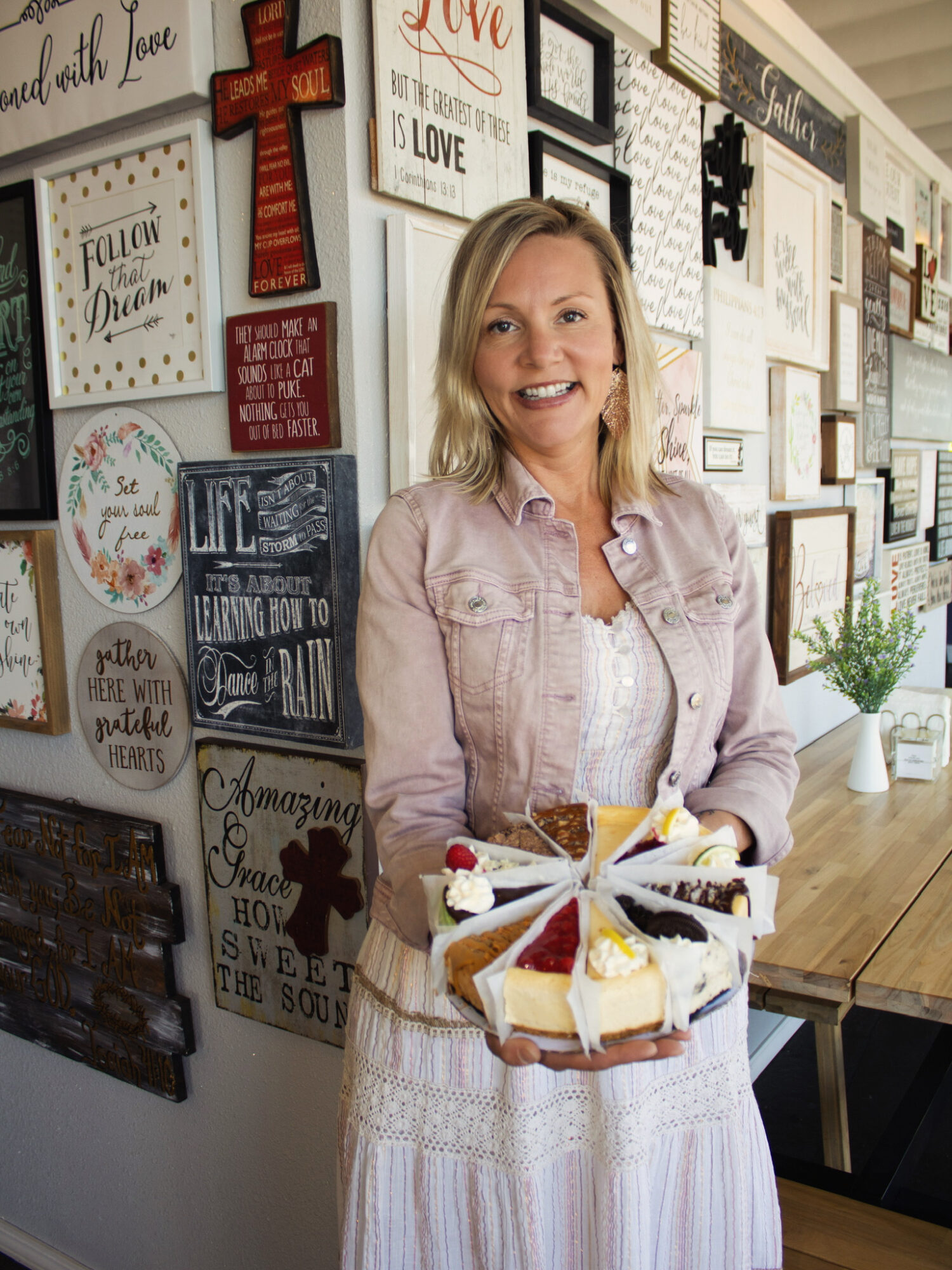Woman with blonde hair smiling, holding a cake with strawberries and cream, standing in front of a wall with framed quotes.