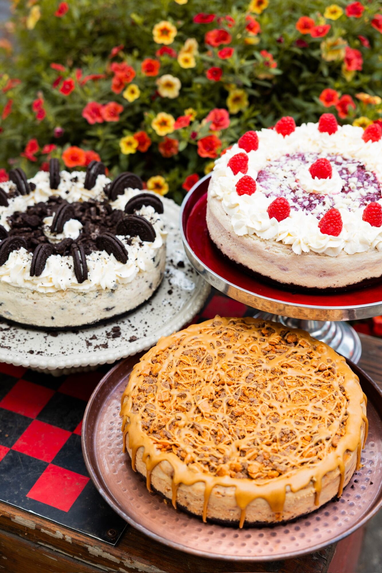 Three cakes on a table with colorful flowers in the background, including a spider cake, a berry-topped cake, and a caramel cake.