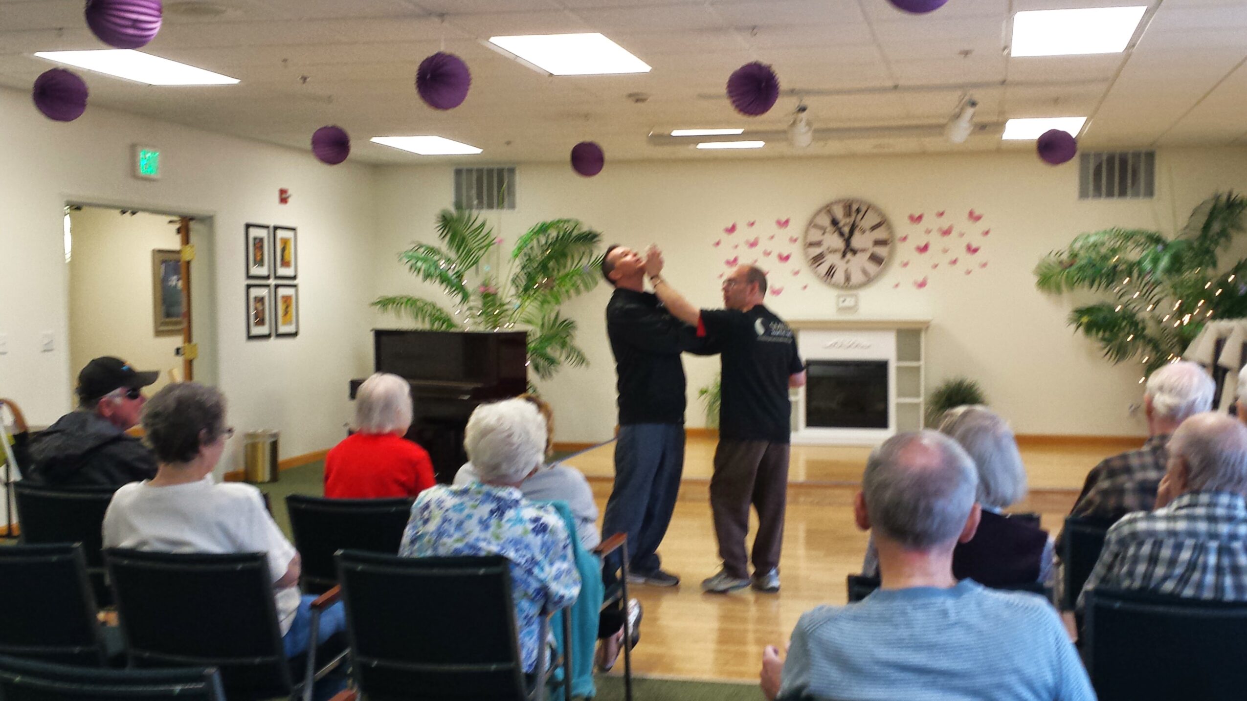 Two performers dance in front of seated audience in a room with plants and a clock, purple decorations hang from ceiling.