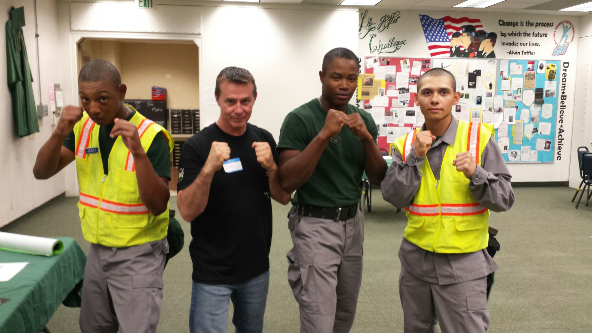Four people standing in a classroom, two wearing yellow safety vests, posing with fists raised.