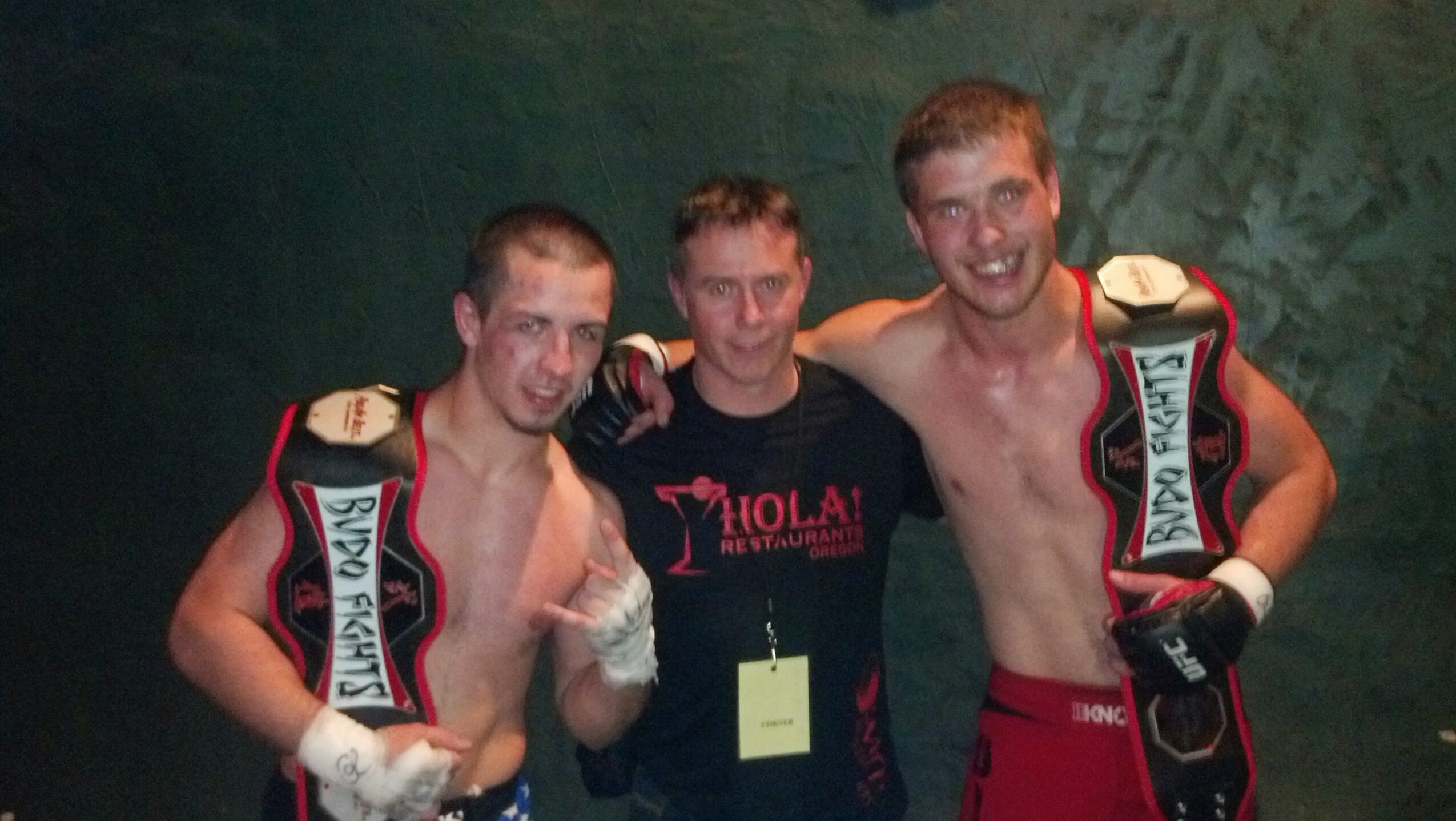 Three men, two shirtless with championship belts, standing together smiling, one in a black shirt, against a green background.