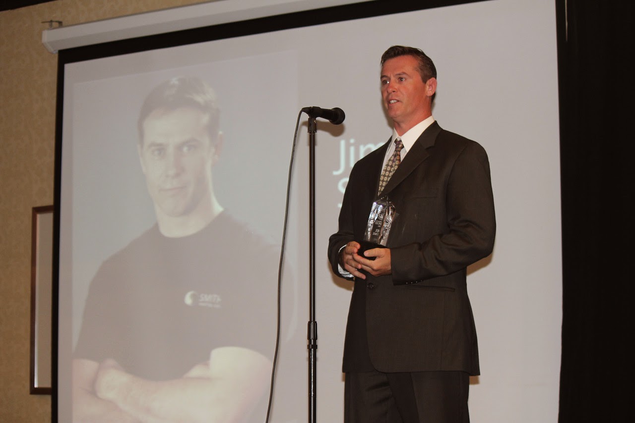Man in suit holding an award, standing in front of a microphone on a stage, with a large screen behind him showing his image.