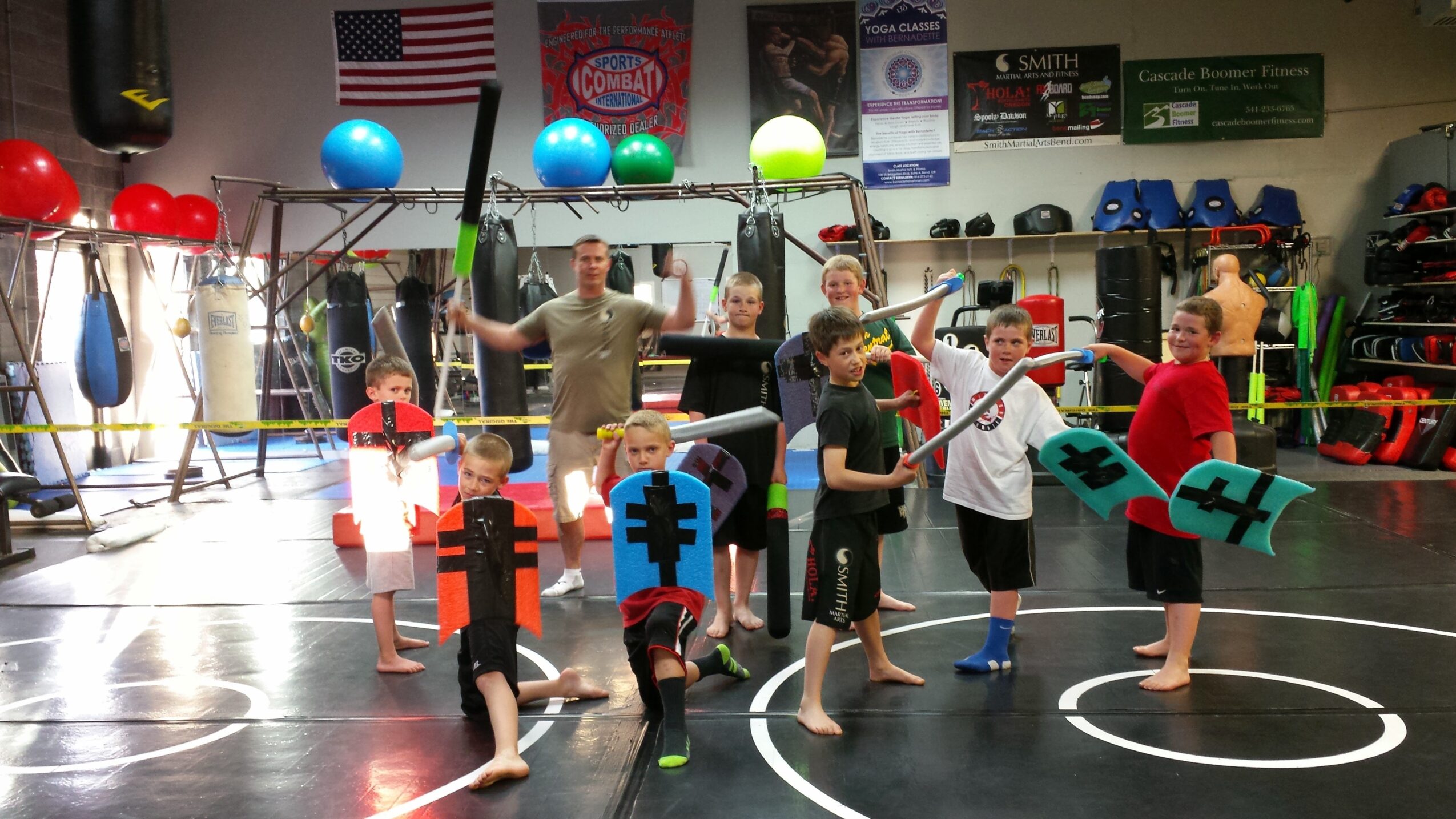 Children and instructor in a martial arts class holding foam swords and shields in a gym.