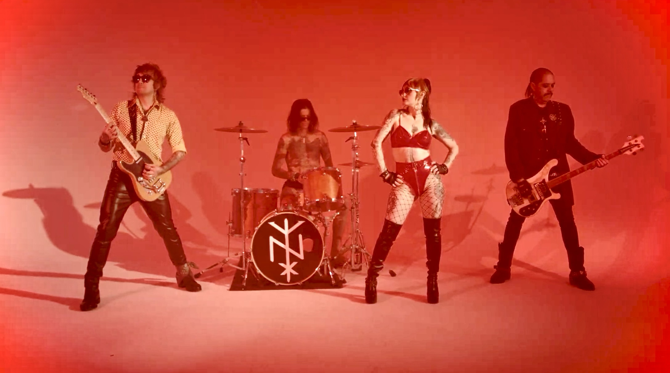 Four musicians perform with guitars and drums under red stage lighting.