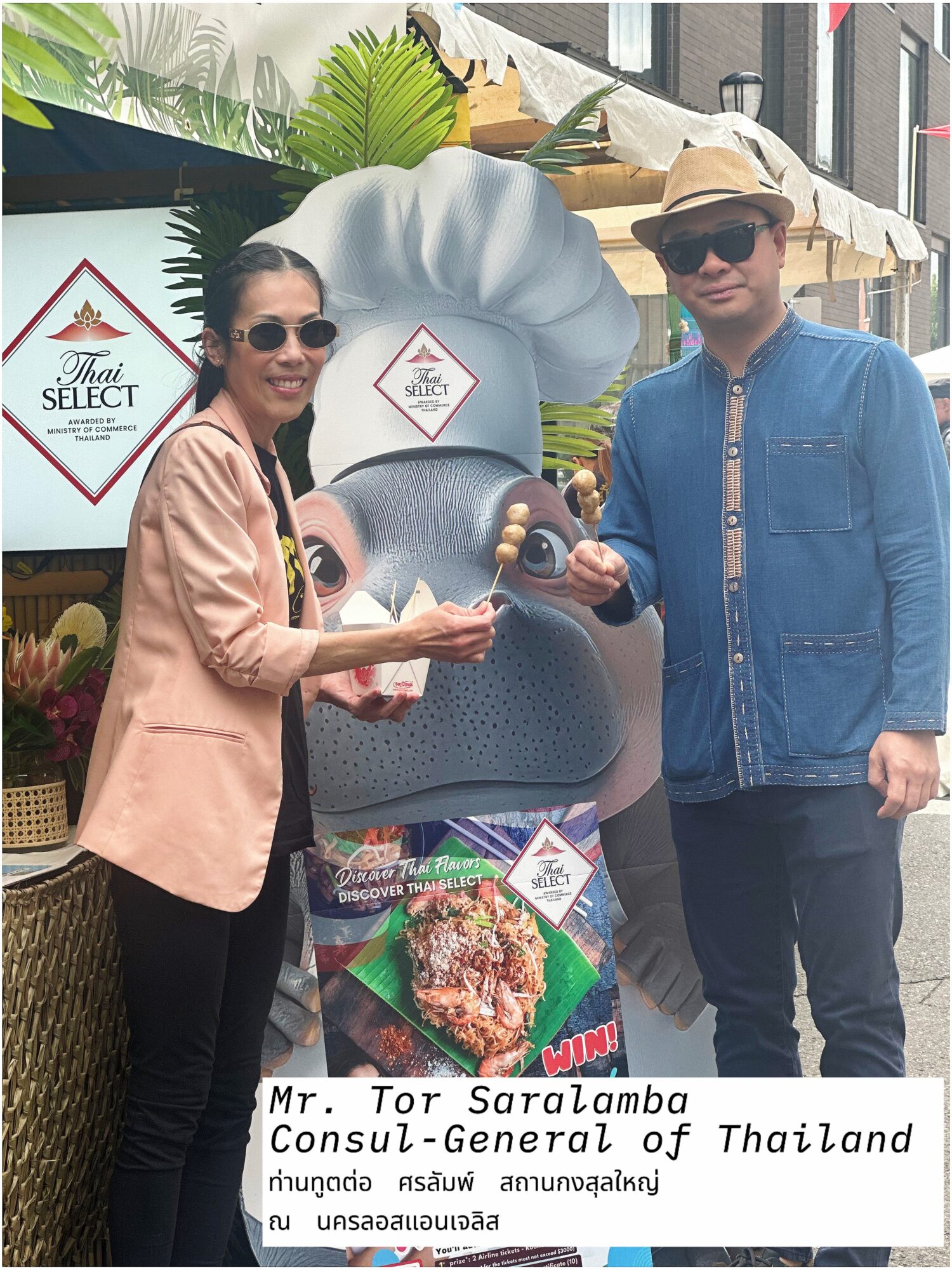 Two people stand beside a large elephant display outside a restaurant, with signs and a food photograph visible.