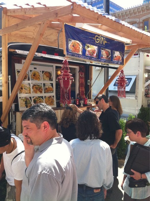 People stand at a wooden food stall with a menu sign and food display.