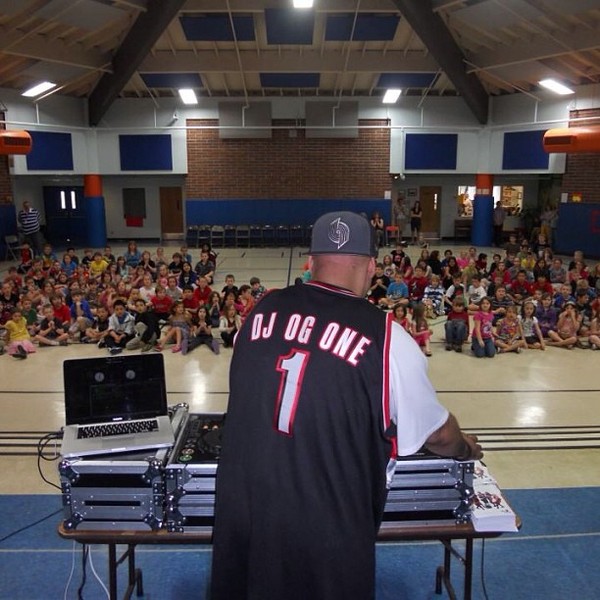 DJ wearing a black and red jersey with the number 1, performing at a school gymnasium with a large audience.