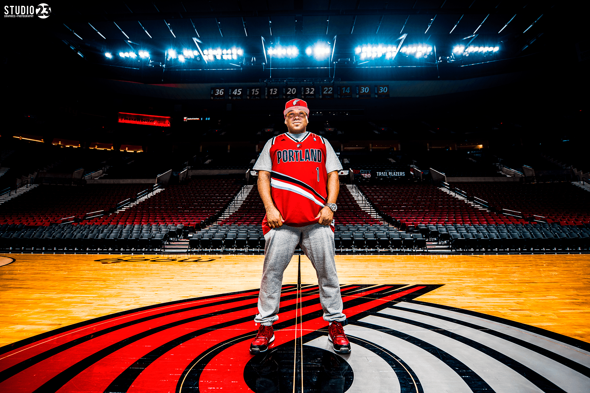 Person standing on basketball court with arena seating and bright lights above, wearing a red and blue sports jersey and cap.