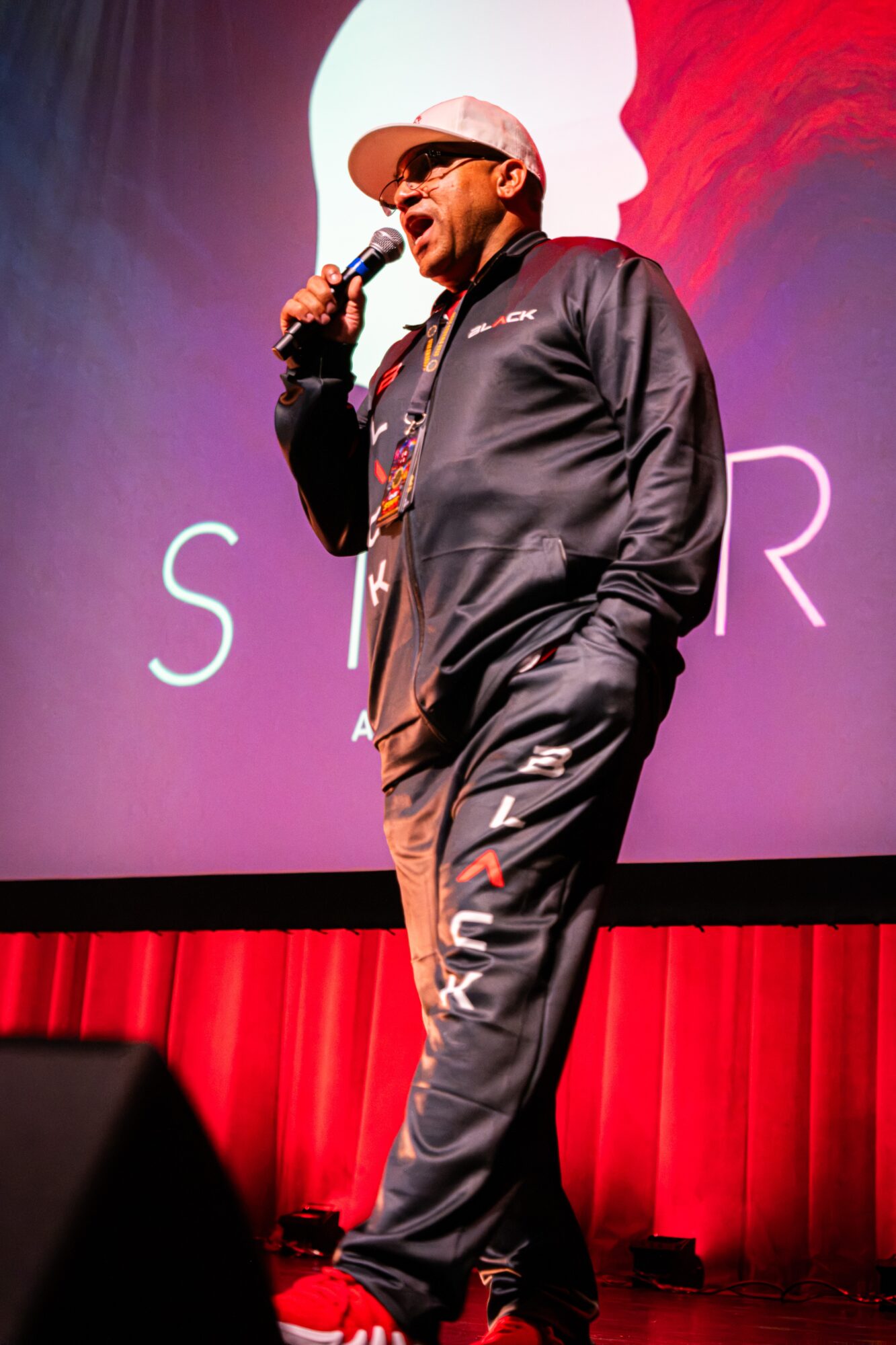 Man in black tracksuit with white cap speaking into microphone on stage with red curtain background.
