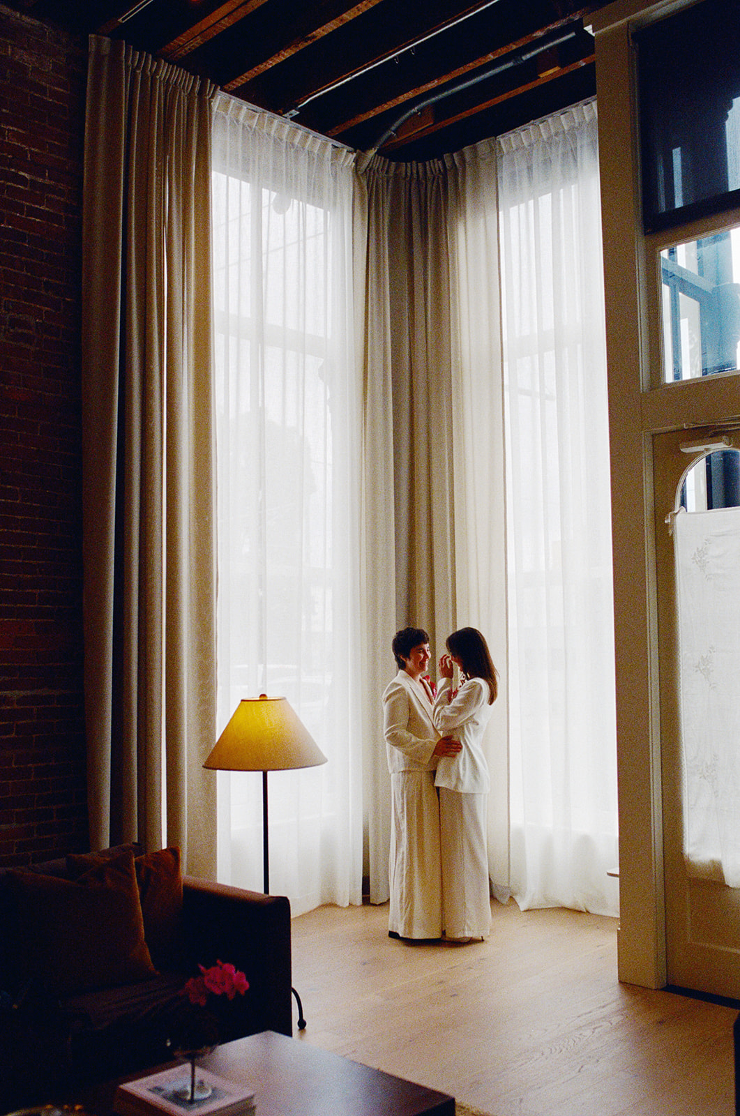 Two people stand close together near large windows with white curtains, in a room with wooden floors and a yellow lamp.