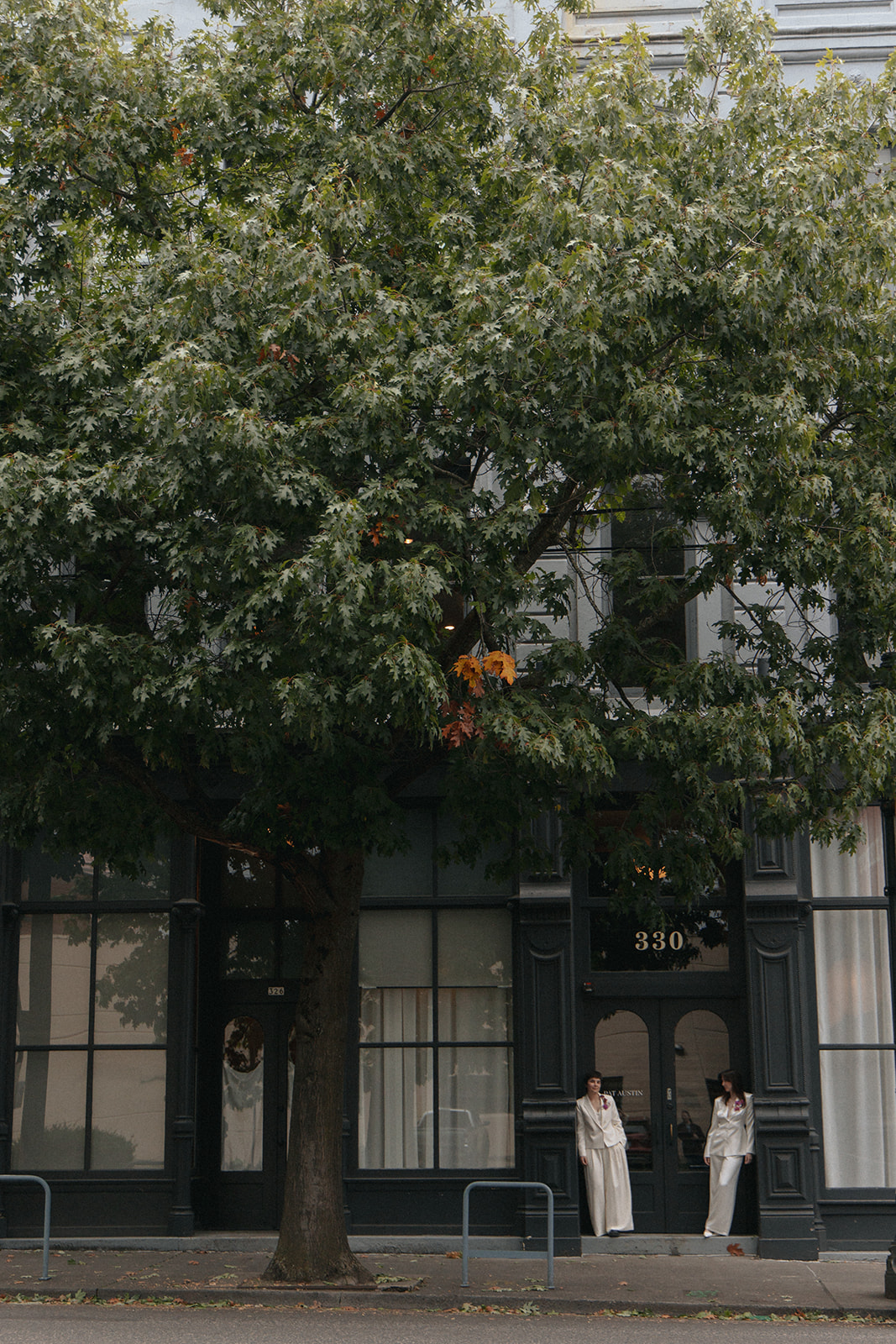 Two people in white outfits stand on sidewalk in front of a building with large windows and a tree with green leaves