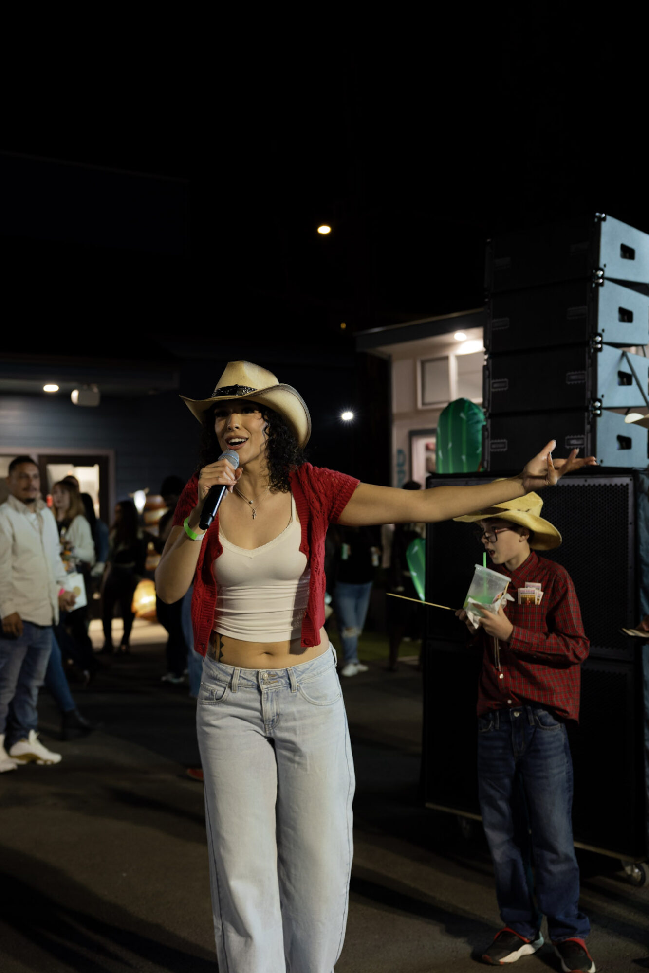 Woman in cowboy hat and red shirt singing into microphone at night, people in background, outdoor setting