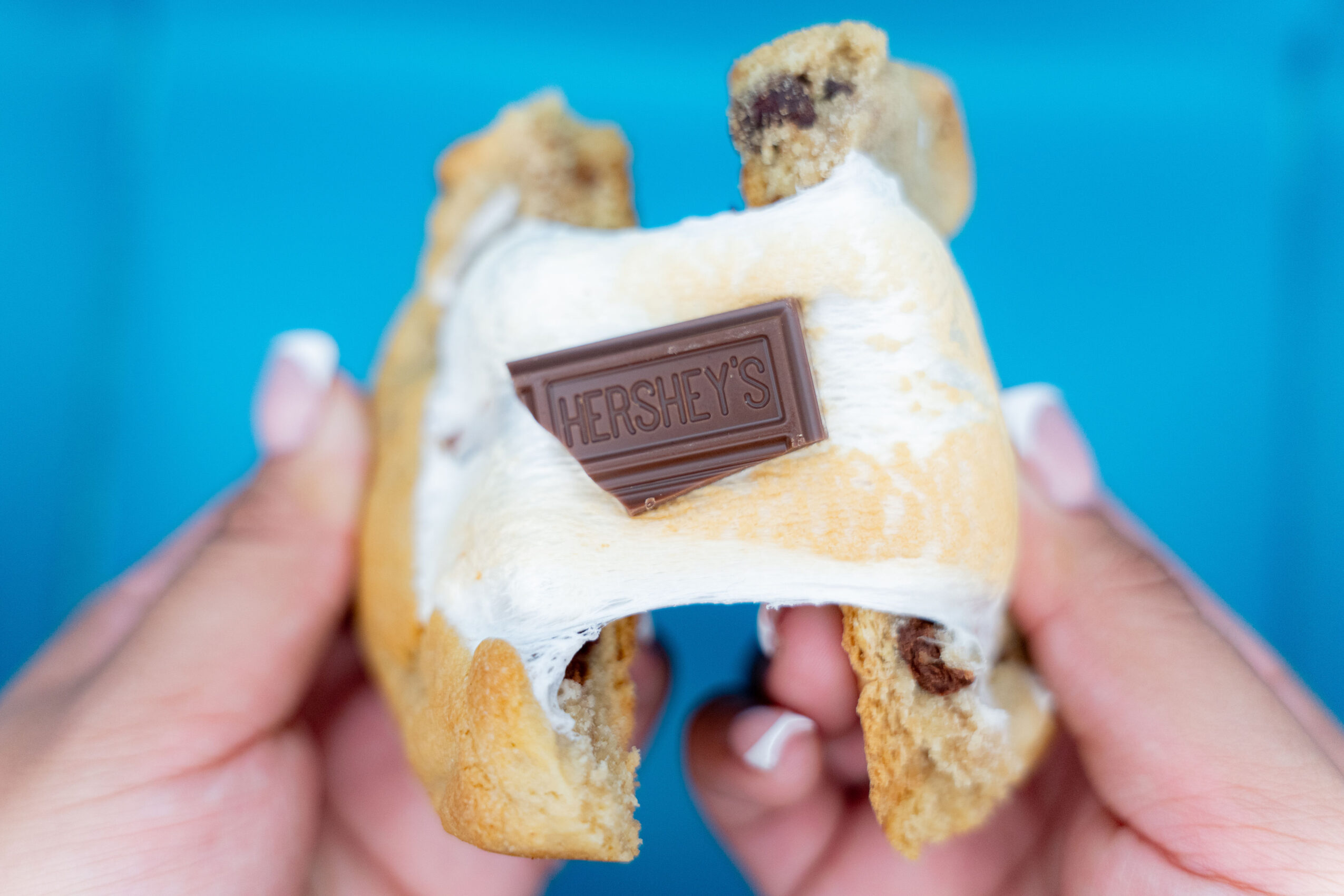 Person holding a partially eaten Hershey's chocolate chip cookie with a piece of chocolate on top, against a blue background.