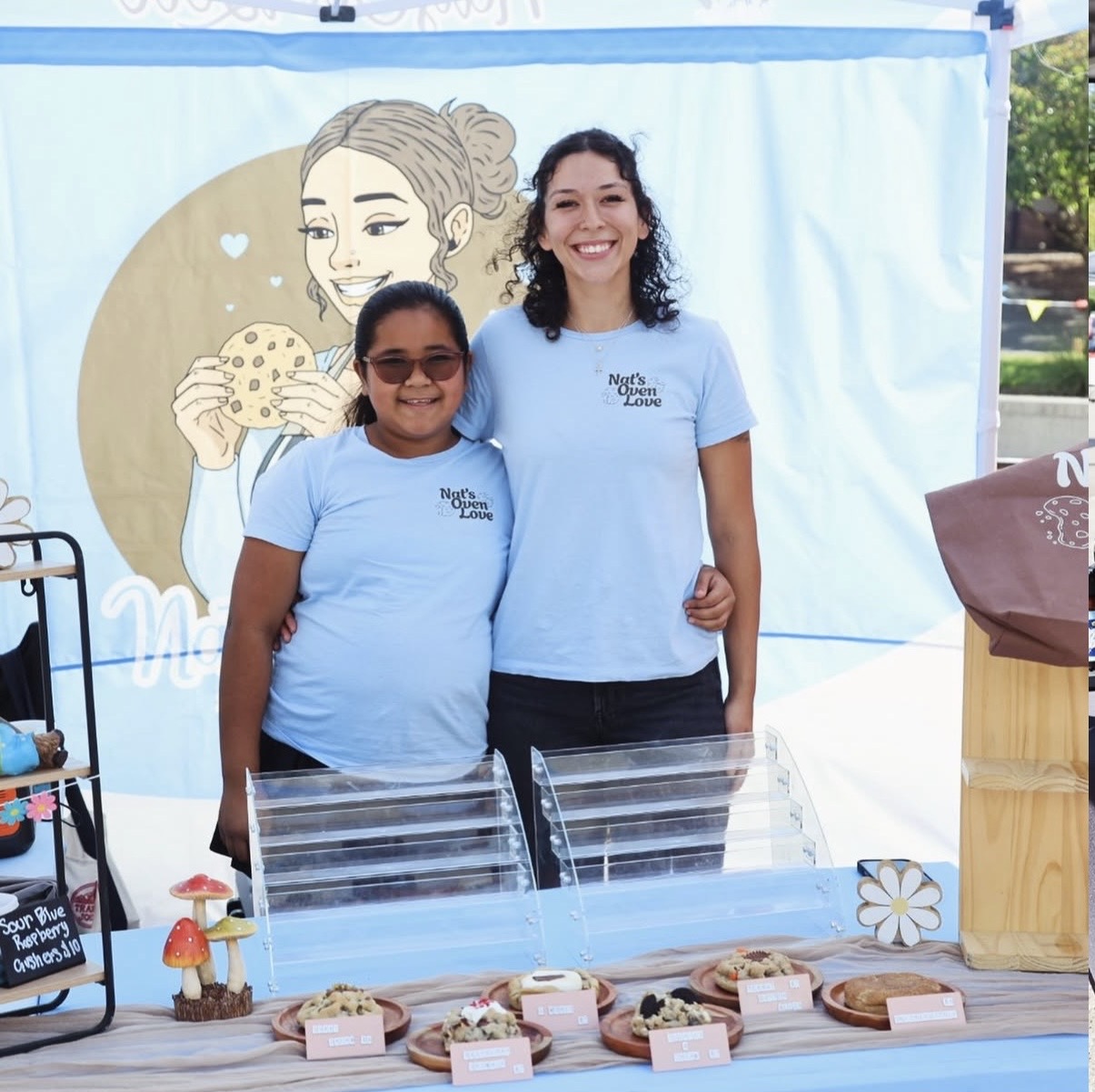 Two smiling women in blue shirts stand behind a table with baked goods, in front of a light blue backdrop with a drawing of a girl eating cookies.