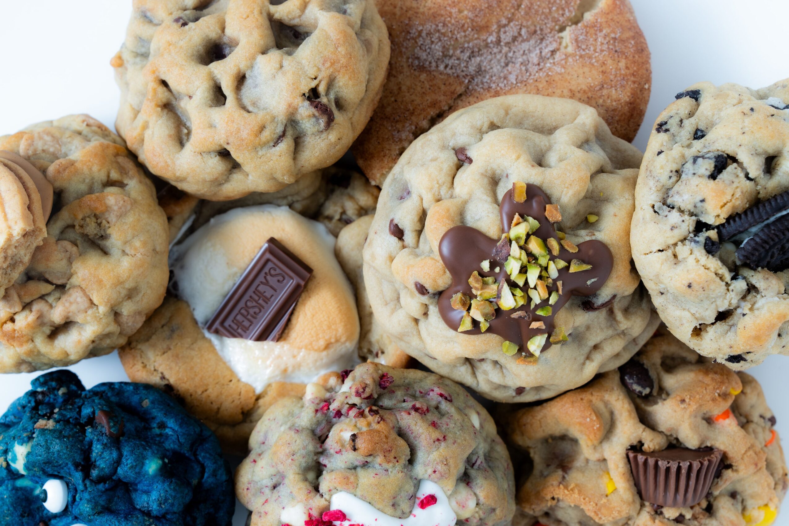 Assorted cookies with chocolate, nuts, and candies, some with chocolate pieces and toppings, on a white surface.