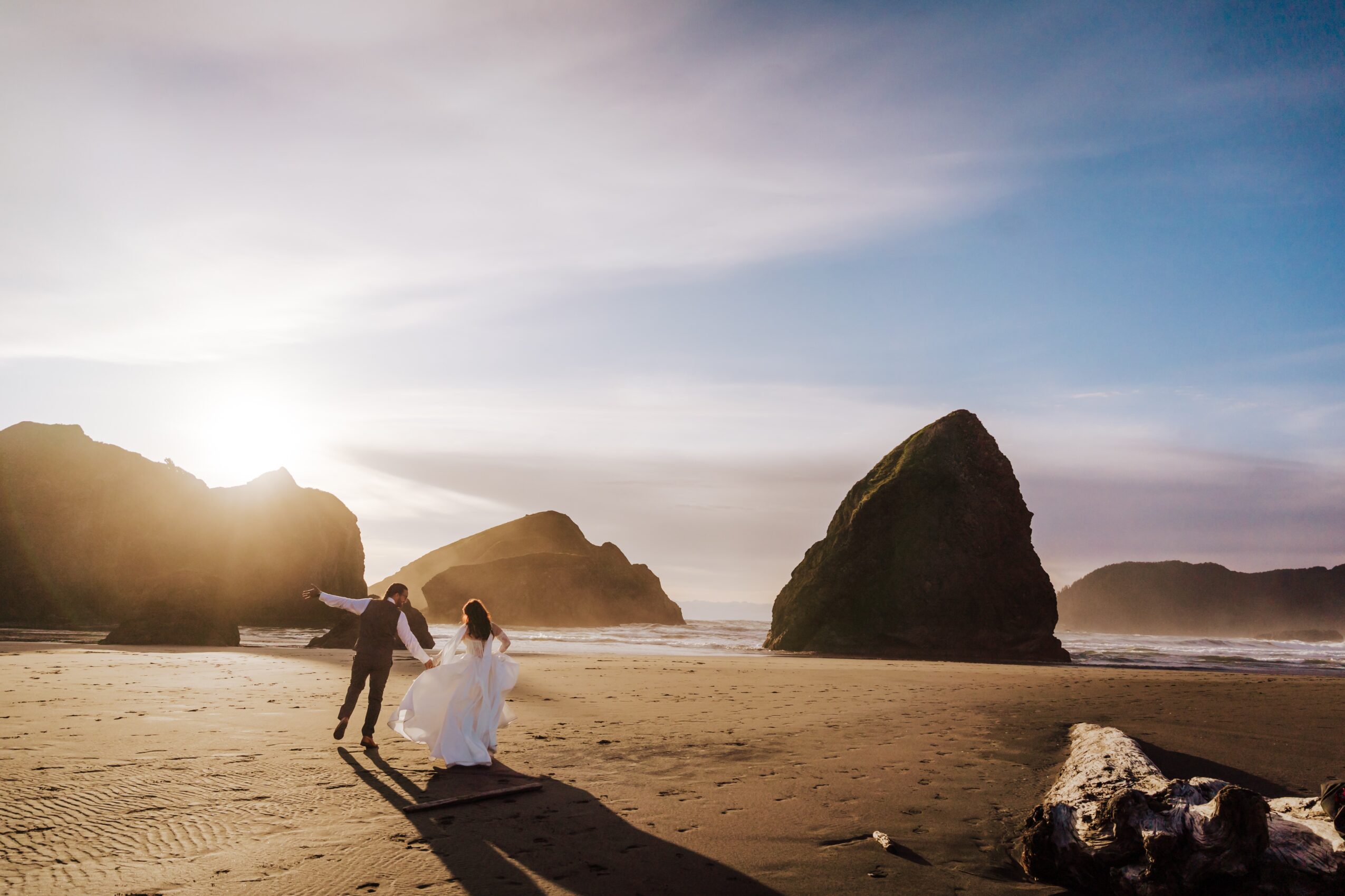 A couple on a beach, with large rocks and ocean waves in the background, during sunset or sunrise, with a partly cloudy sky.