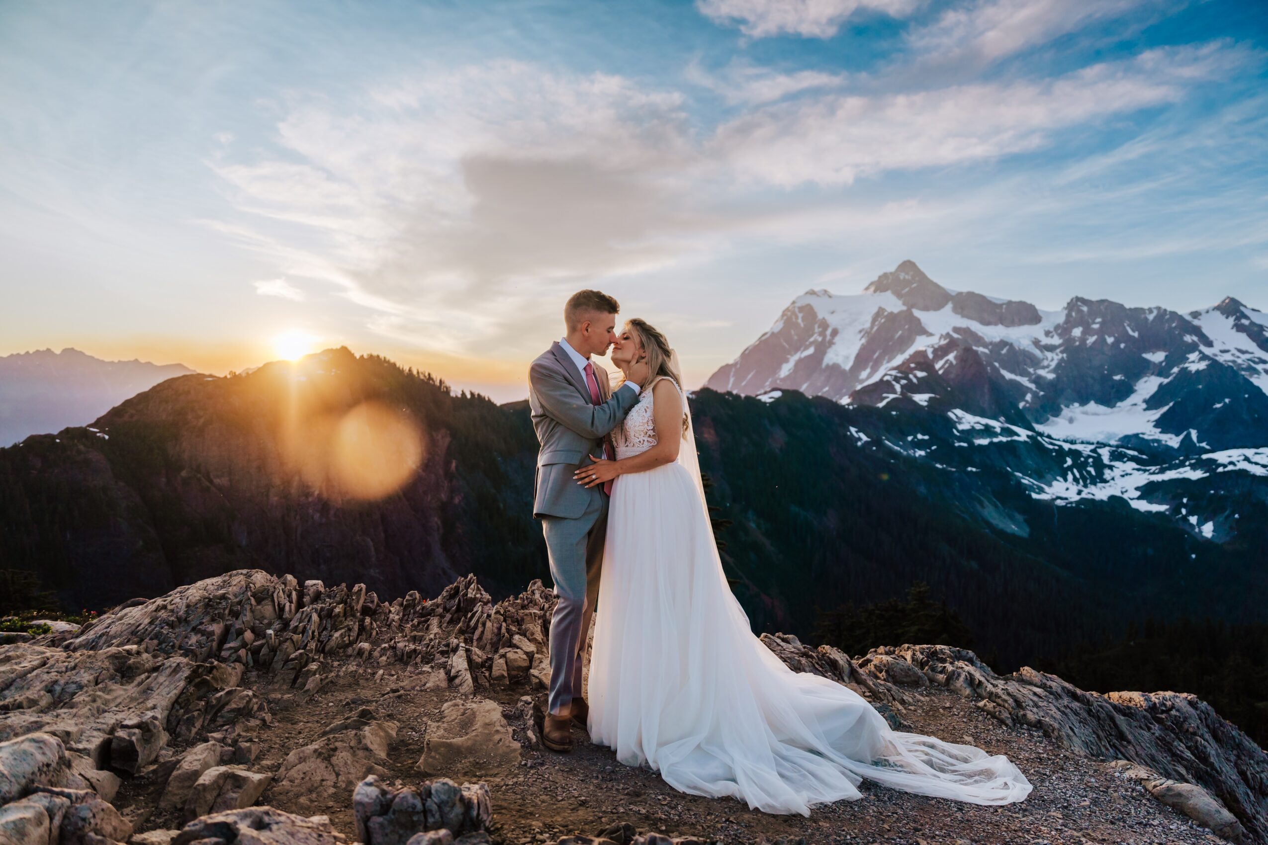 A couple in wedding attire standing on rocky terrain with mountains and a sunset in the background.