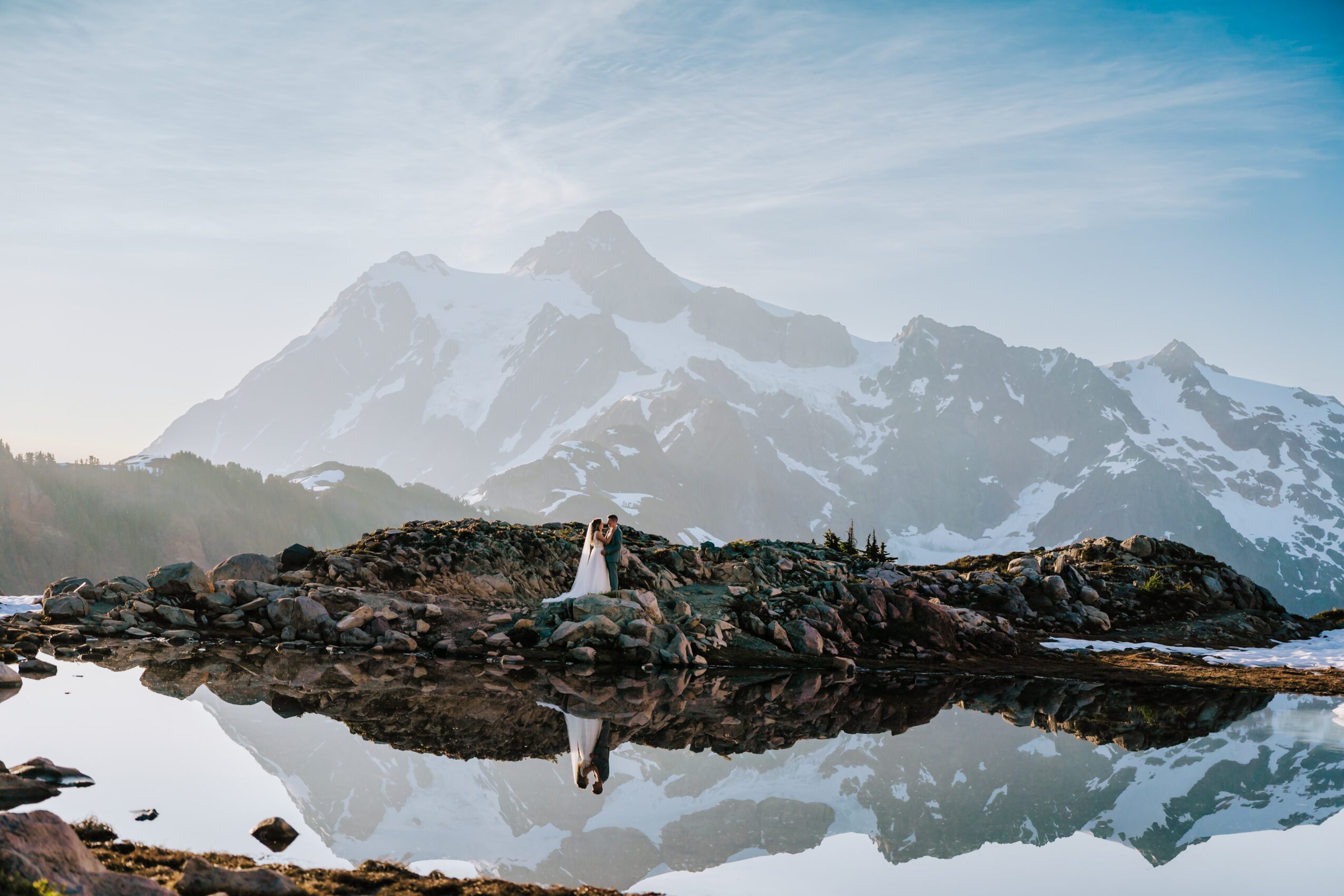 Person standing on rocks near water with snow-capped mountain in background, reflected in water, daytime.
