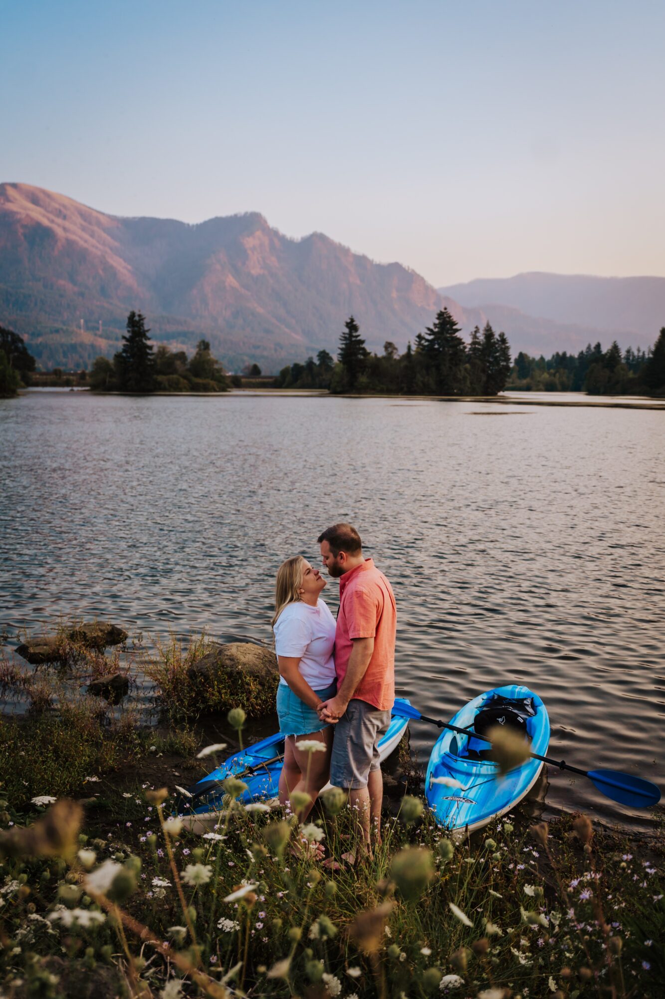 A couple stands by a lake with mountains in the background, holding hands and looking at each other, with a blue kayak nearby.