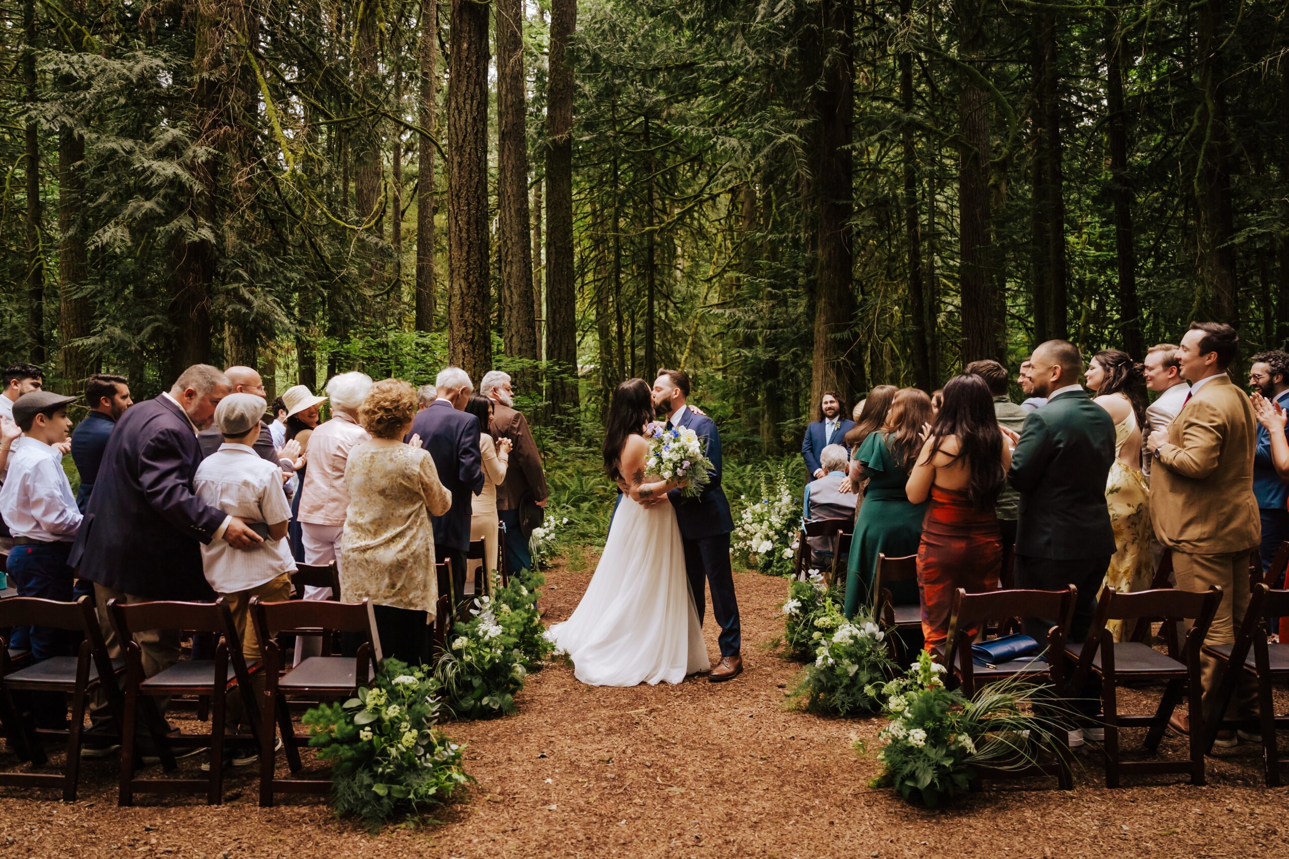 A wedding ceremony in a forest with a bride and groom surrounded by guests, chairs, and greenery.