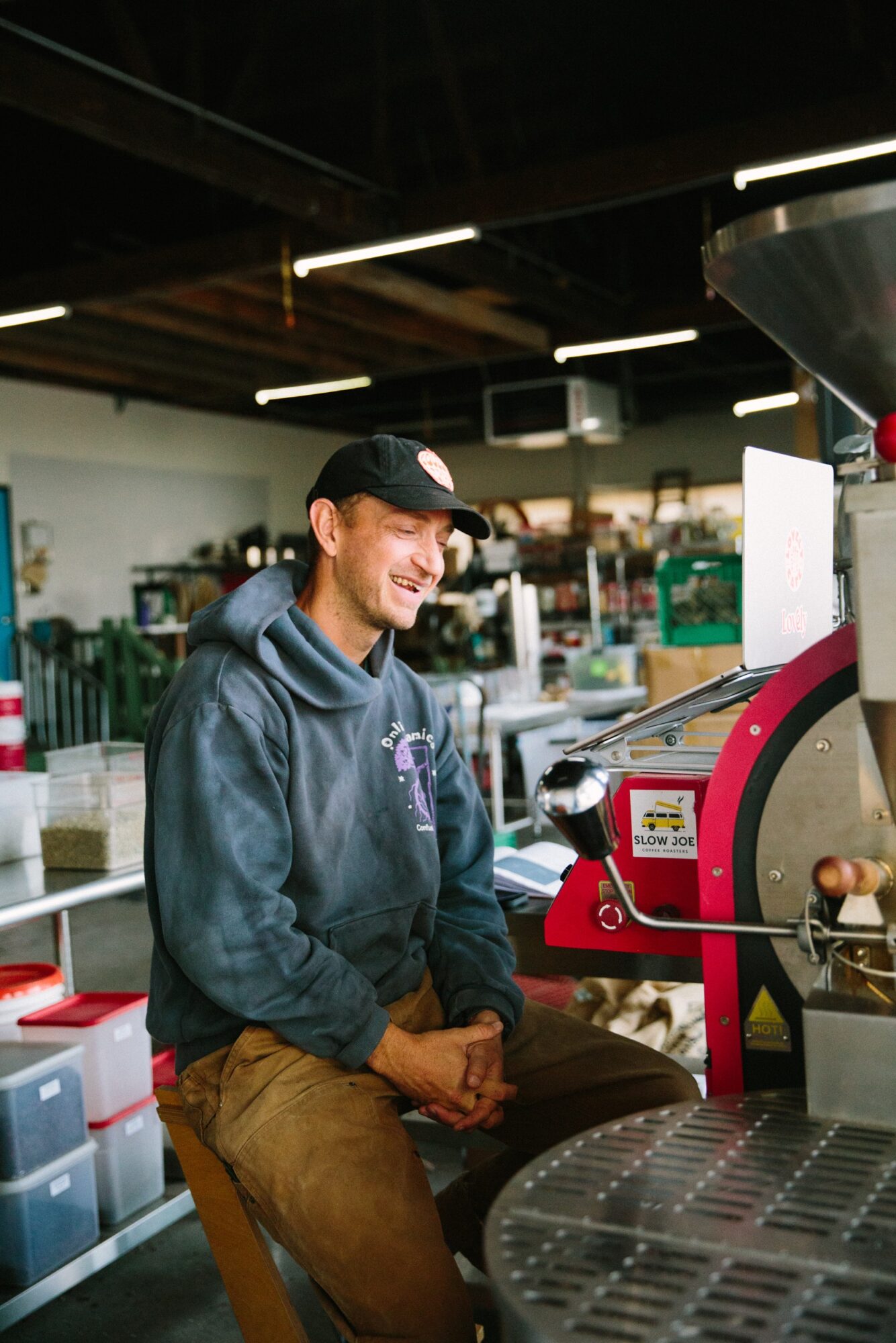 Young man sitting in a coffee shop, smiling, wearing a black cap and hoodie, with coffee equipment in the background.