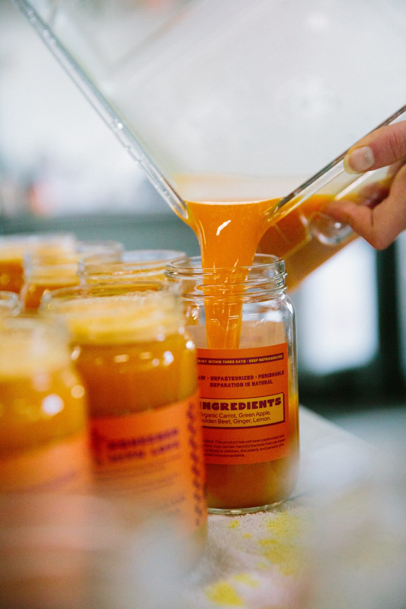 Pouring orange liquid into a jar with a label, with several jars in the foreground, in a bright setting.