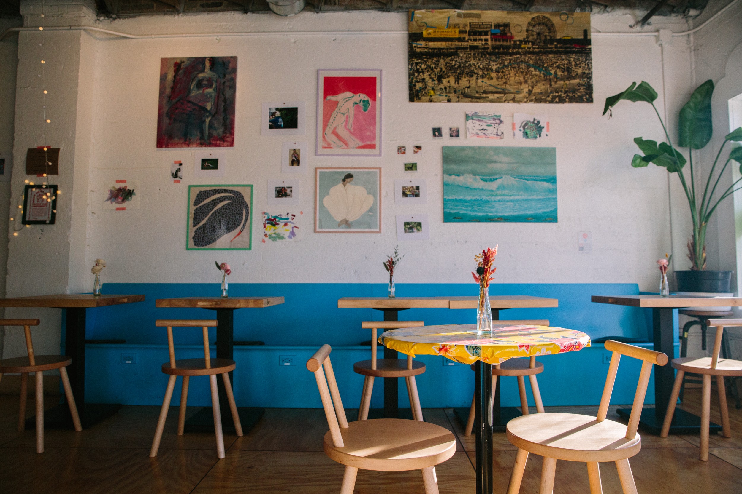 Interior of a cafe with tables, chairs, and artwork on the wall, large plant on the right, and a blue bench.