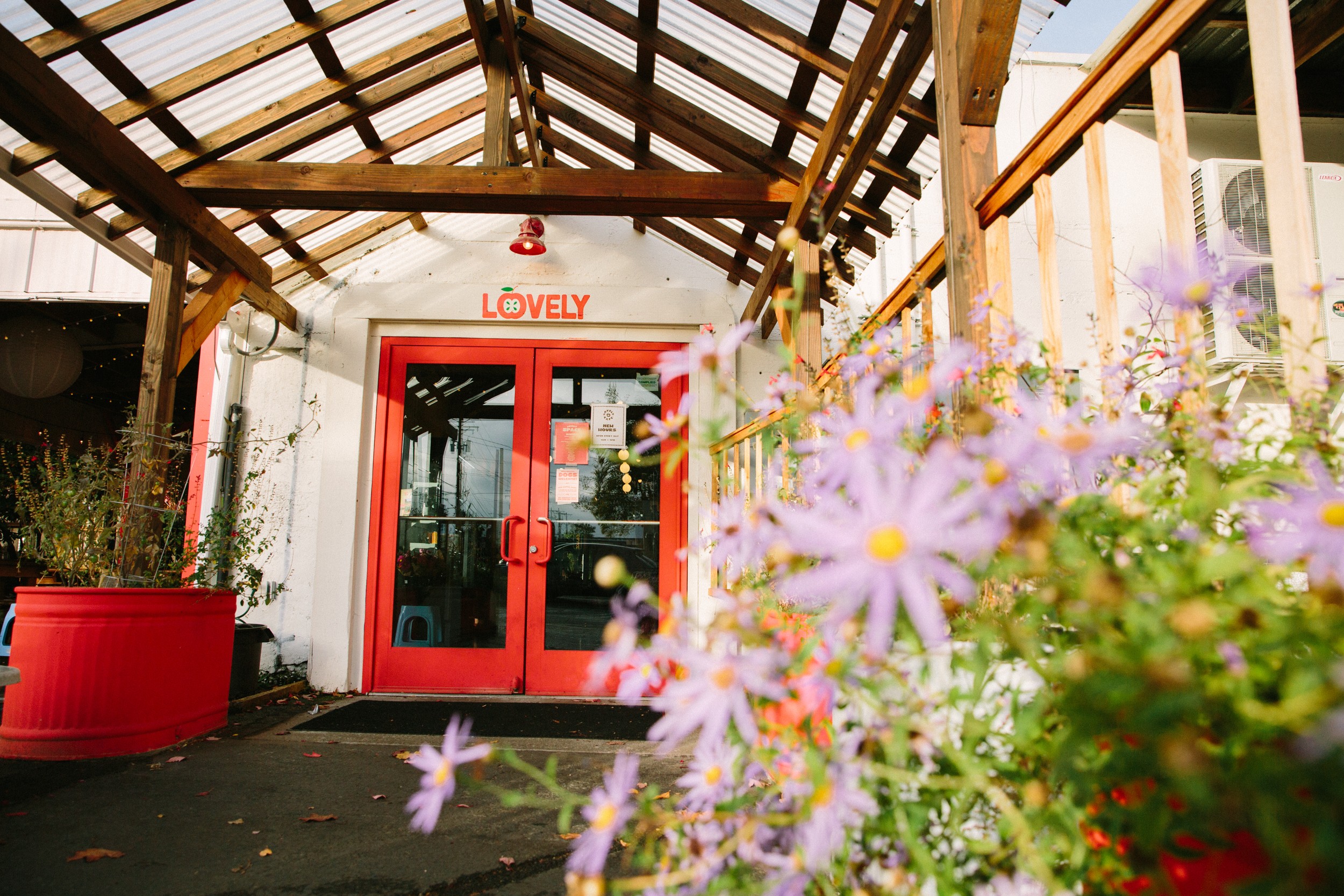 Entrance with red door and glass panels, surrounded by flowers and plants, under a wooden roof structure, bright outdoor setting.