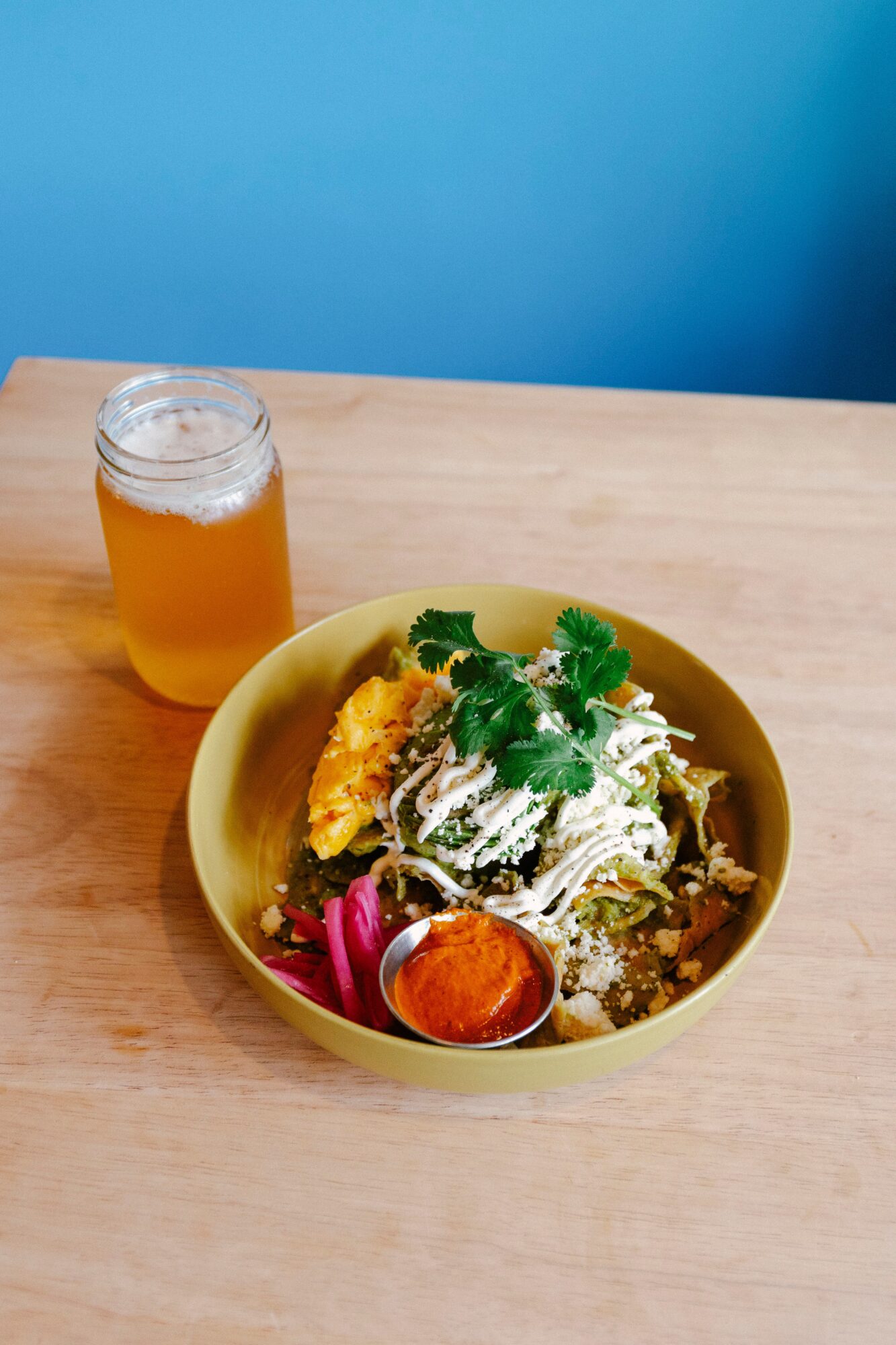Bowl of salad with greens, vegetables, and cheese, next to a jar of orange-colored drink on a wooden table.