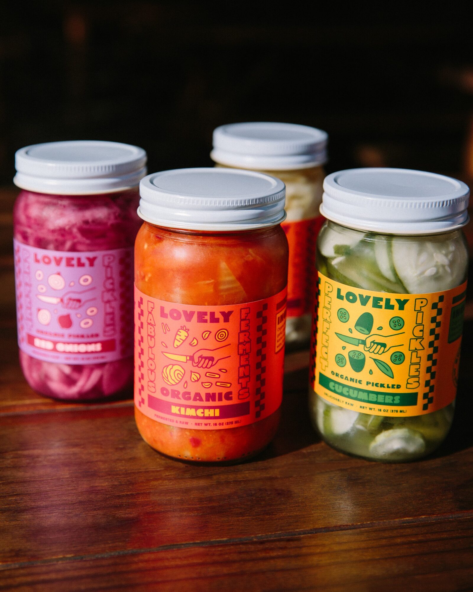 Four jars with colorful labels and white lids on a wooden surface, containing various preserved foods.