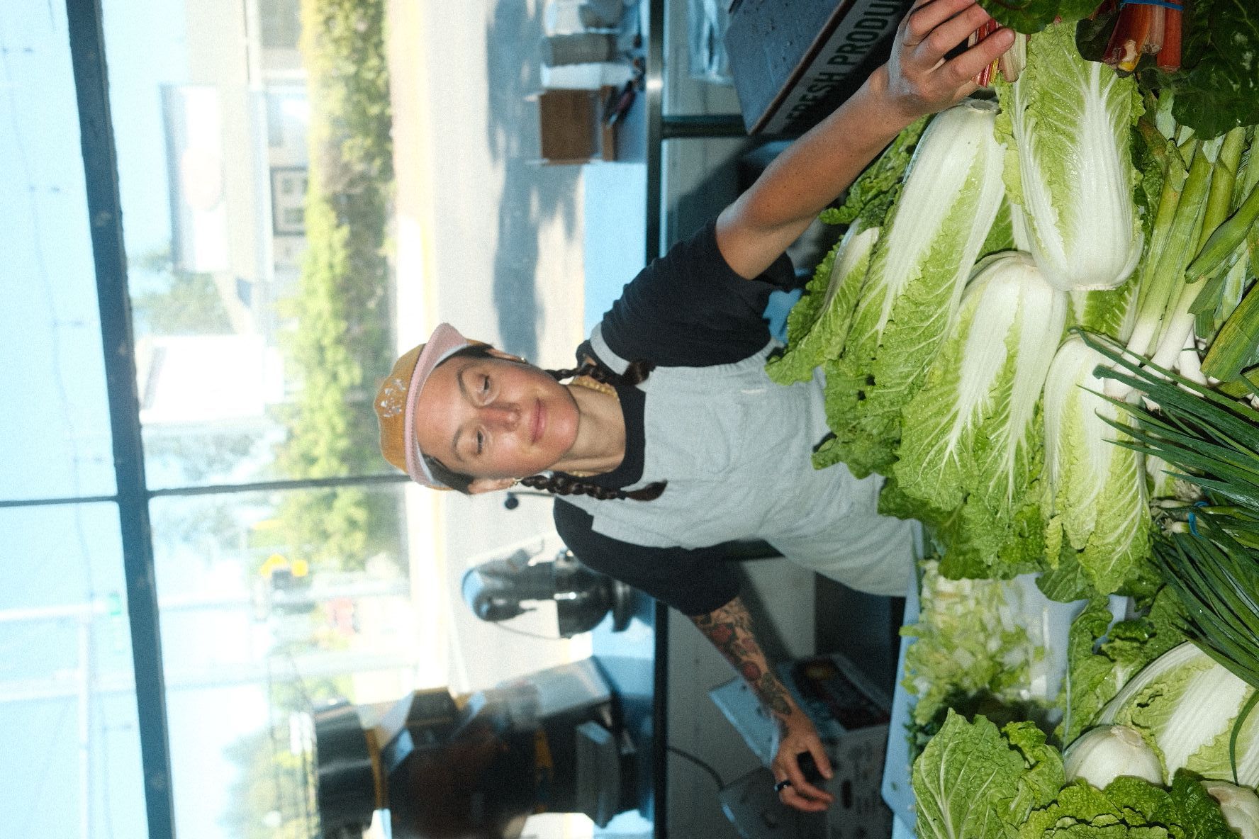 Person with glasses and a white shirt arranging lettuce at a market stall, with produce and a glass window behind.