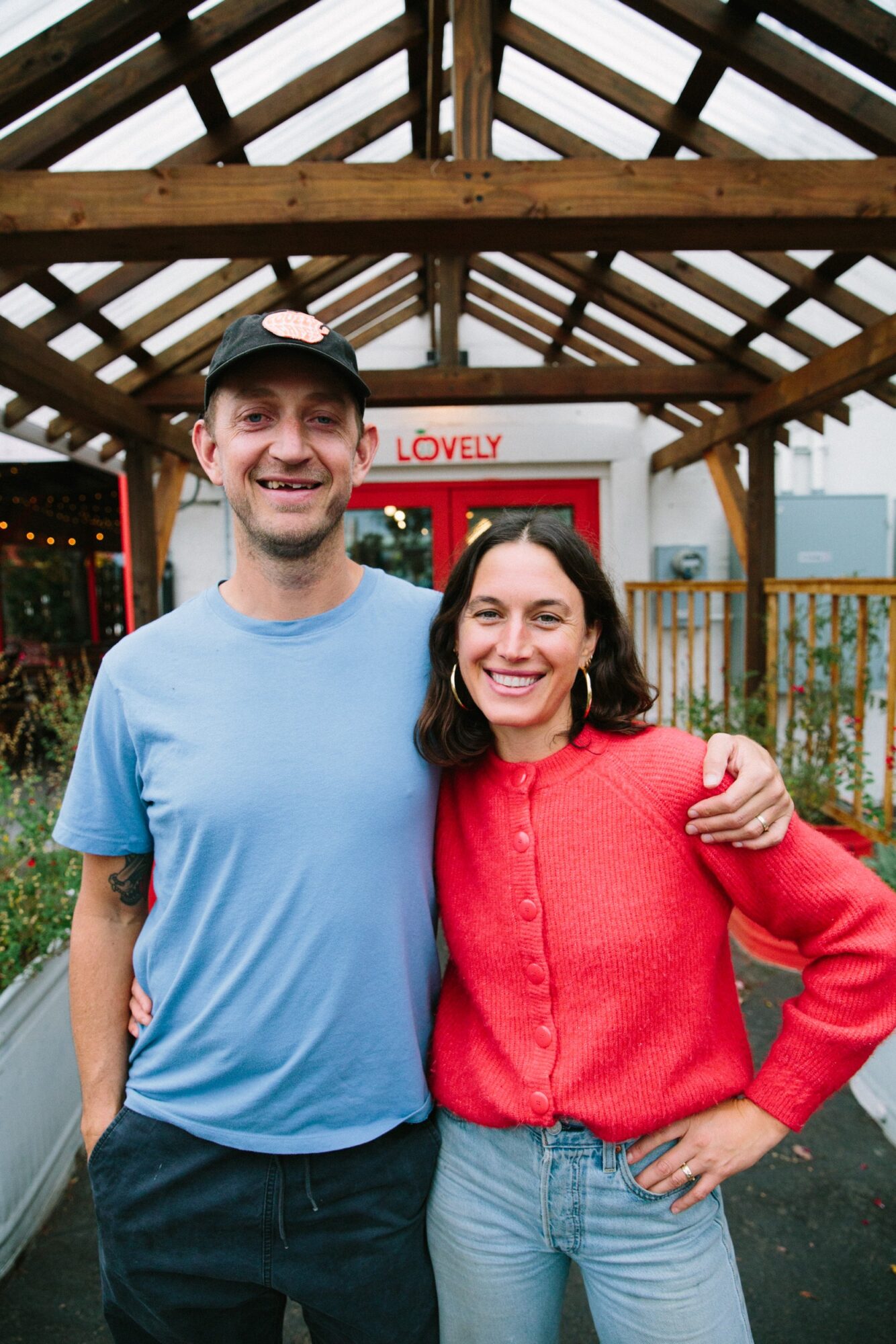 Two people smiling, standing outdoors under a wooden roof structure, with a red and white sign in the background.
