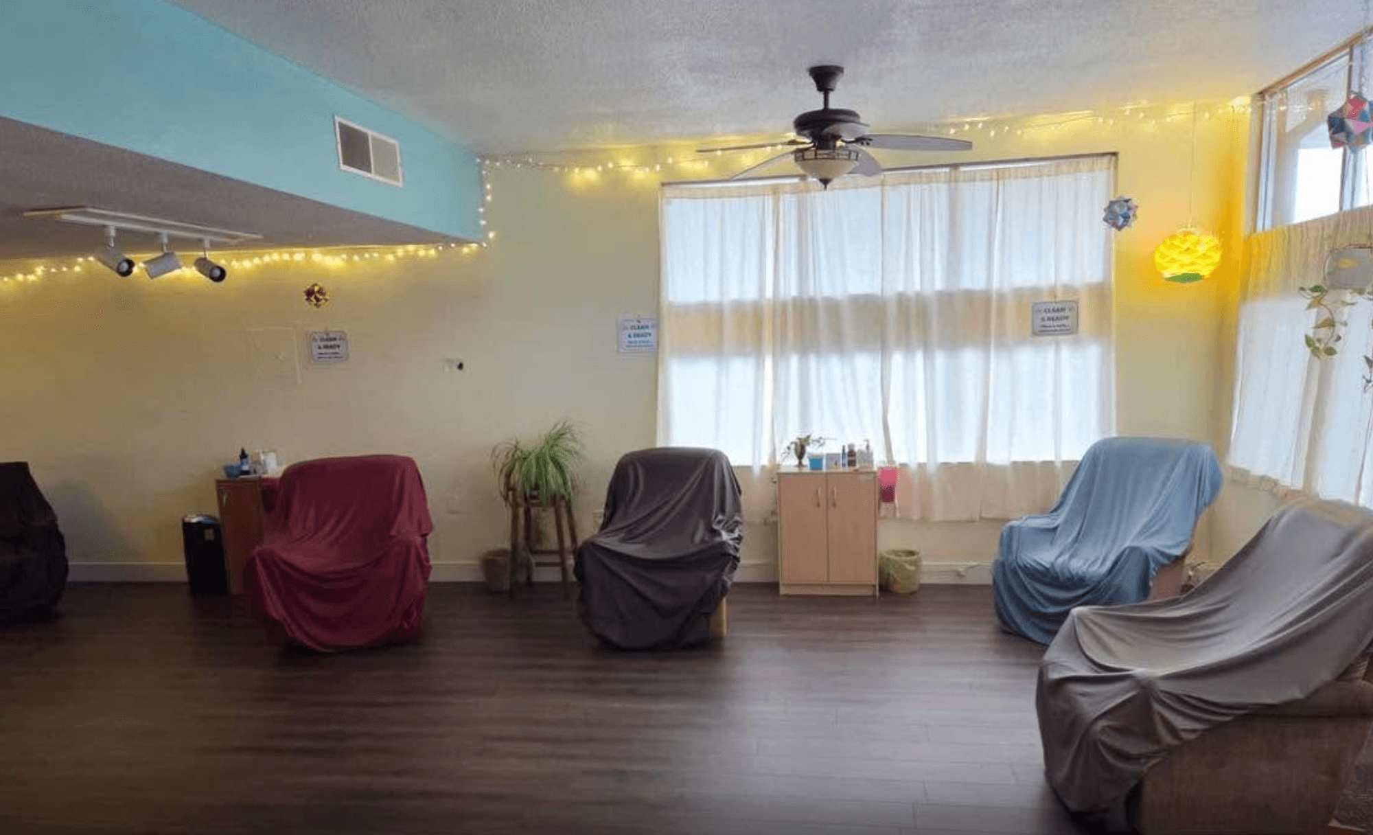A room with curtained windows, a ceiling fan, string lights, and several furniture pieces covered with fabric.