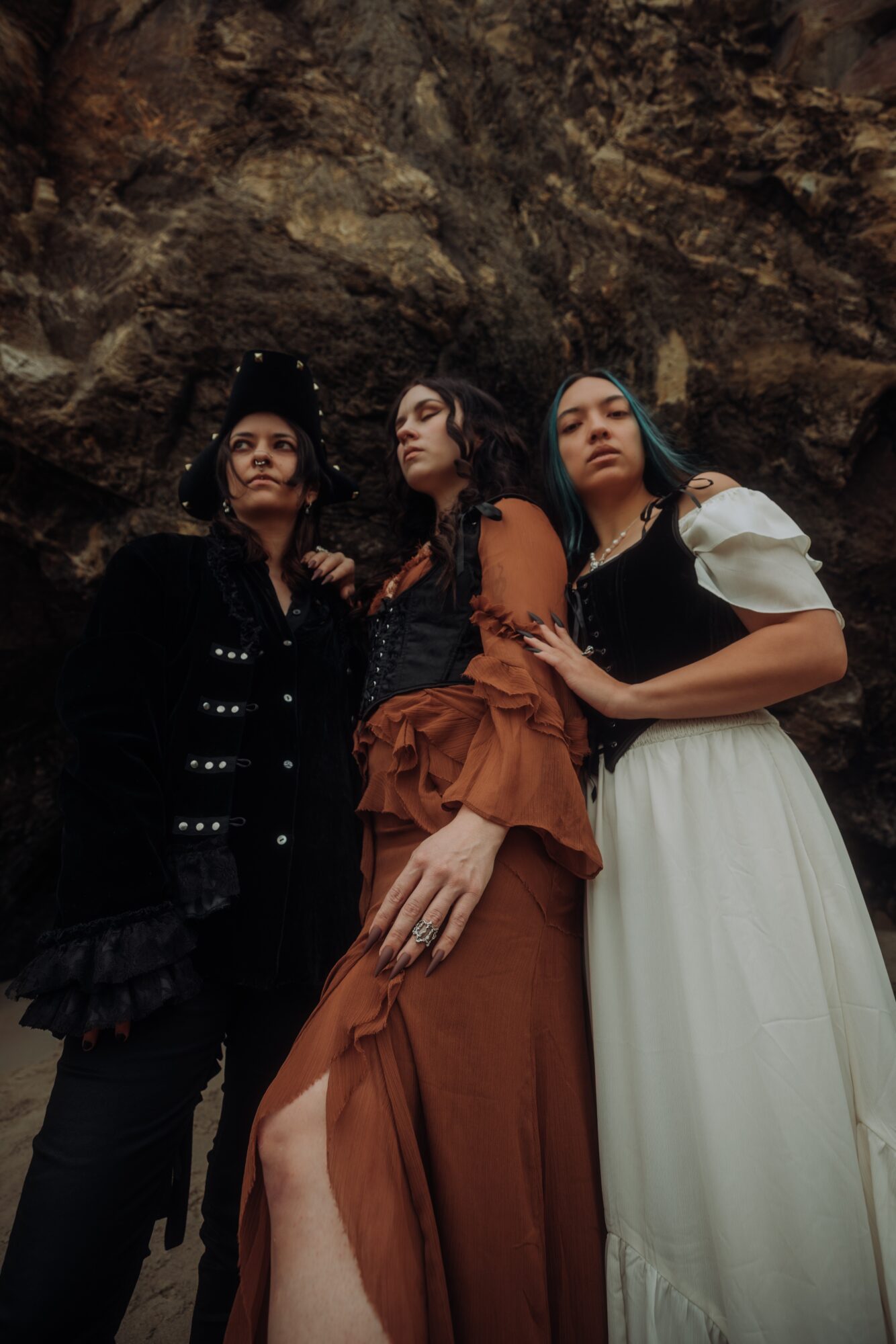 Three women standing close together against a rocky background, dressed in dark and white clothing, with serious expressions.