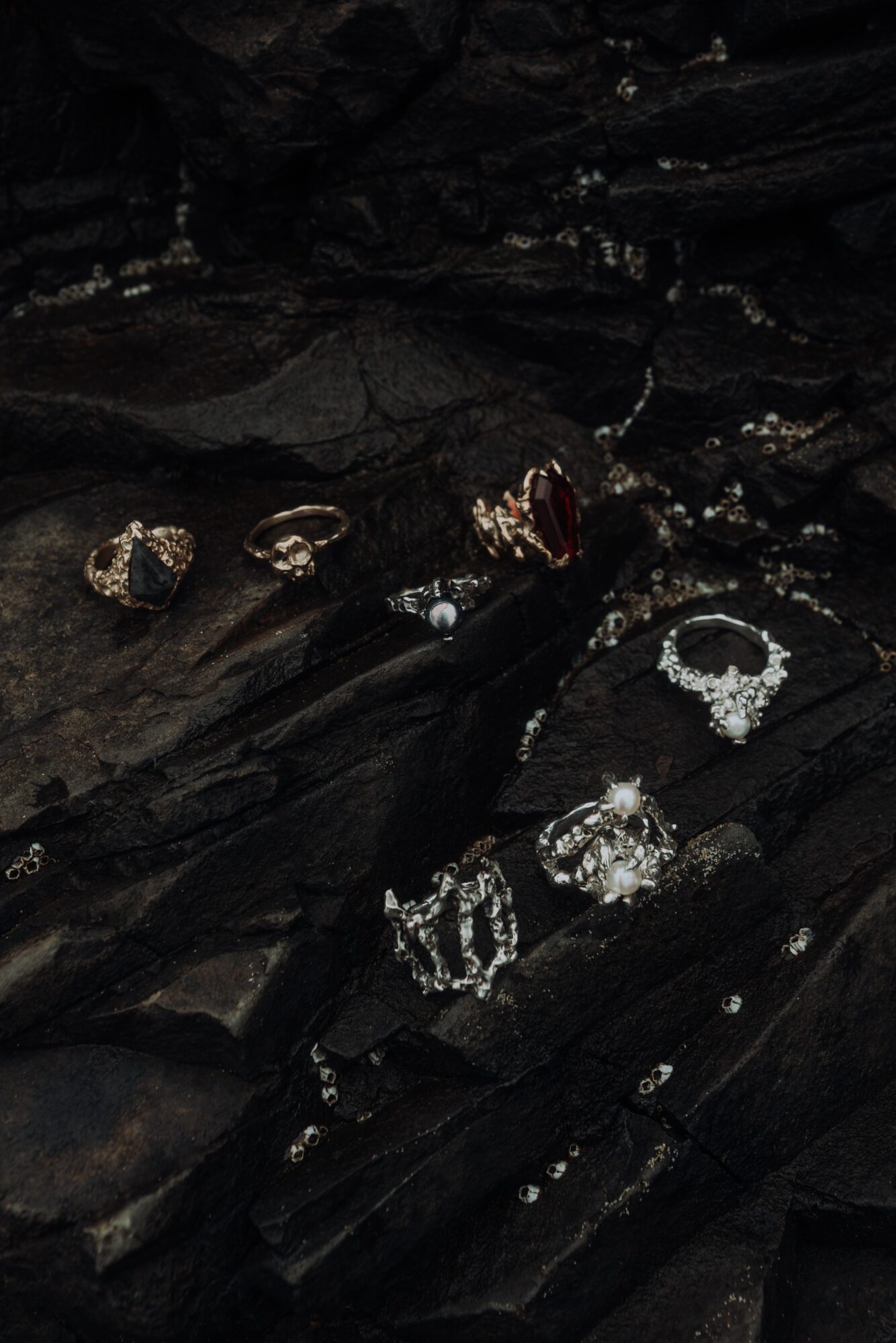 Multiple rings and jewelry pieces placed on dark, textured rocks, with some shiny and reflective surfaces.