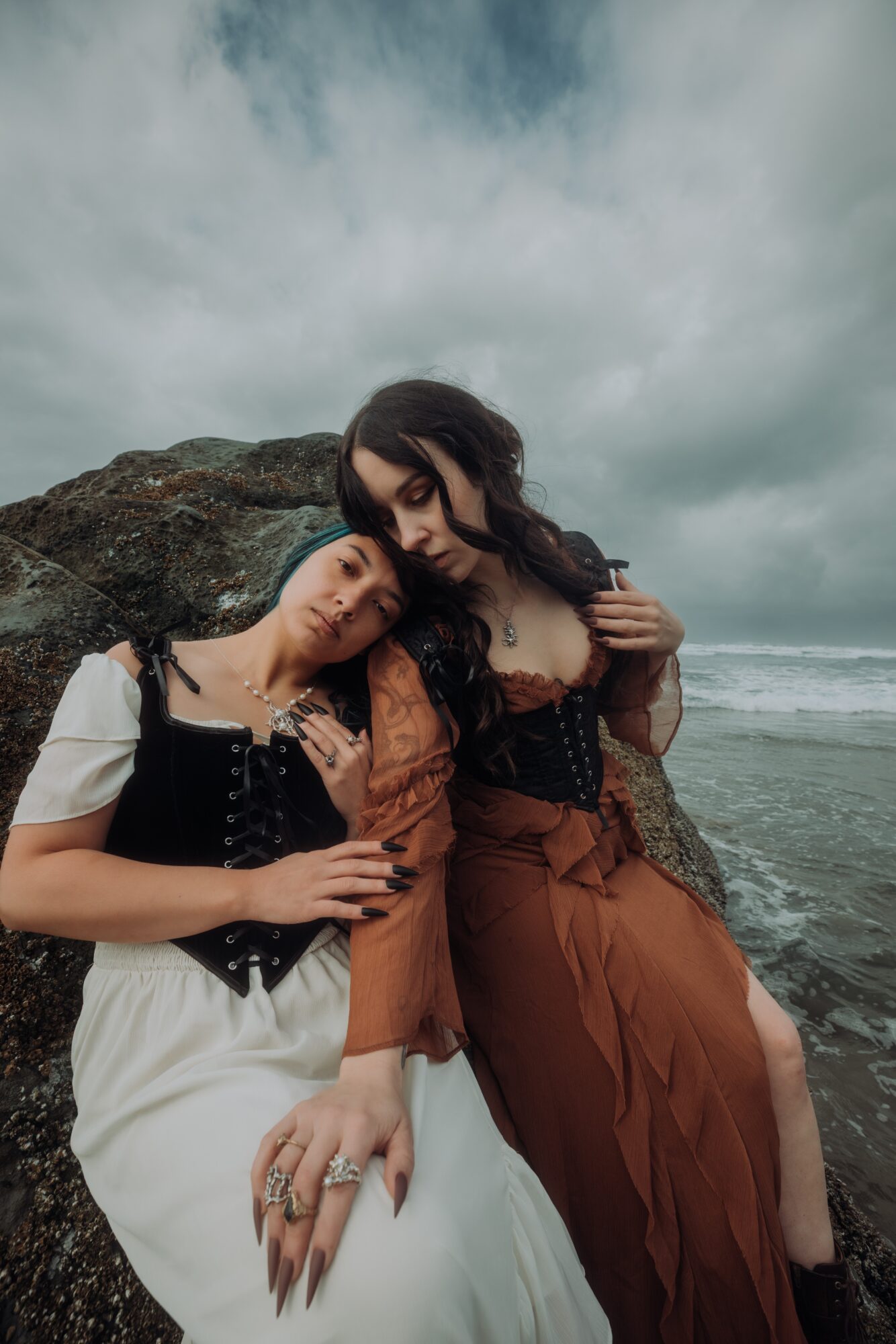 Two women sit on a large rock at the beach, with cloudy sky and ocean in the background.