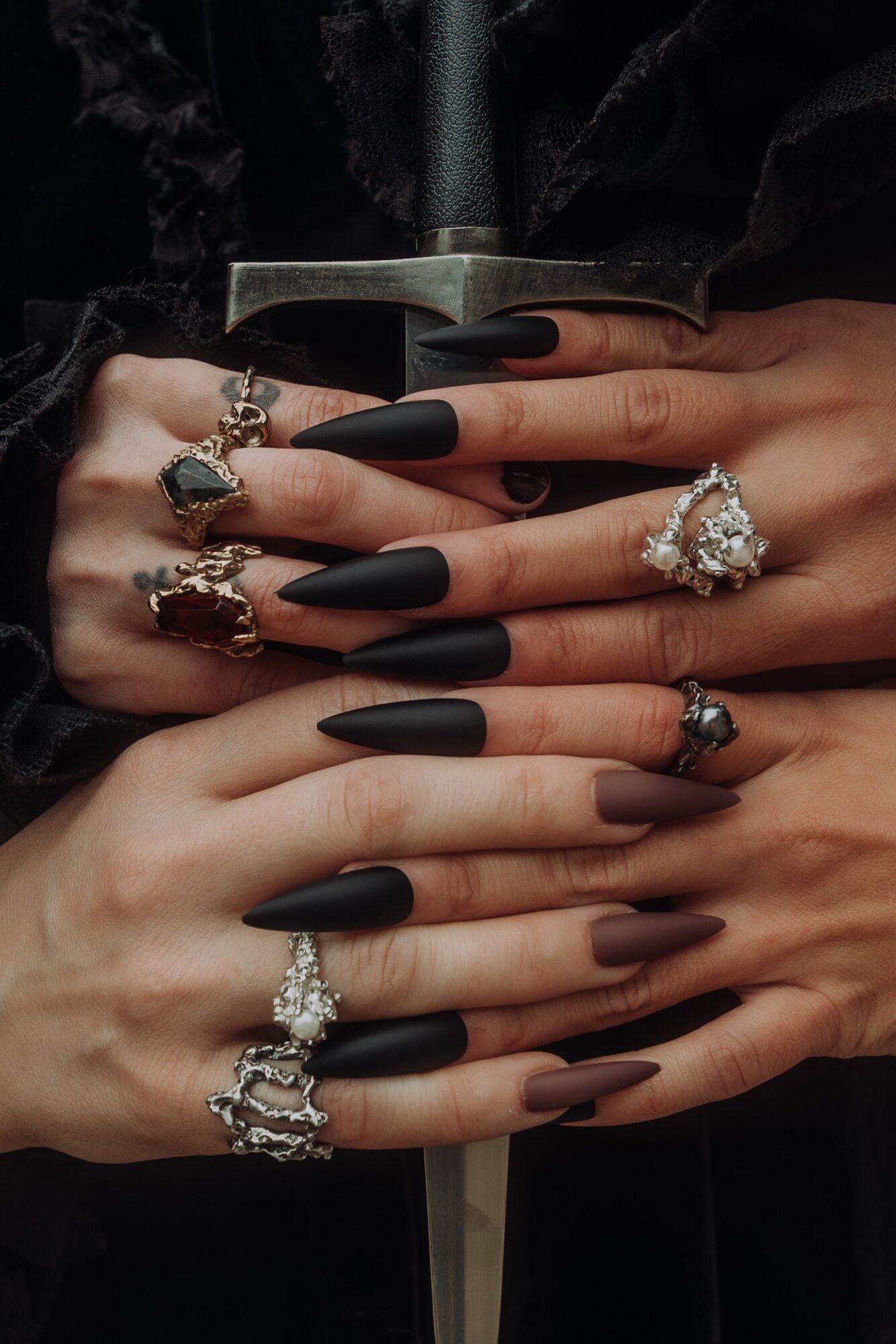 Multiple hands with rings and black matte nails, holding a book, against dark clothing background.